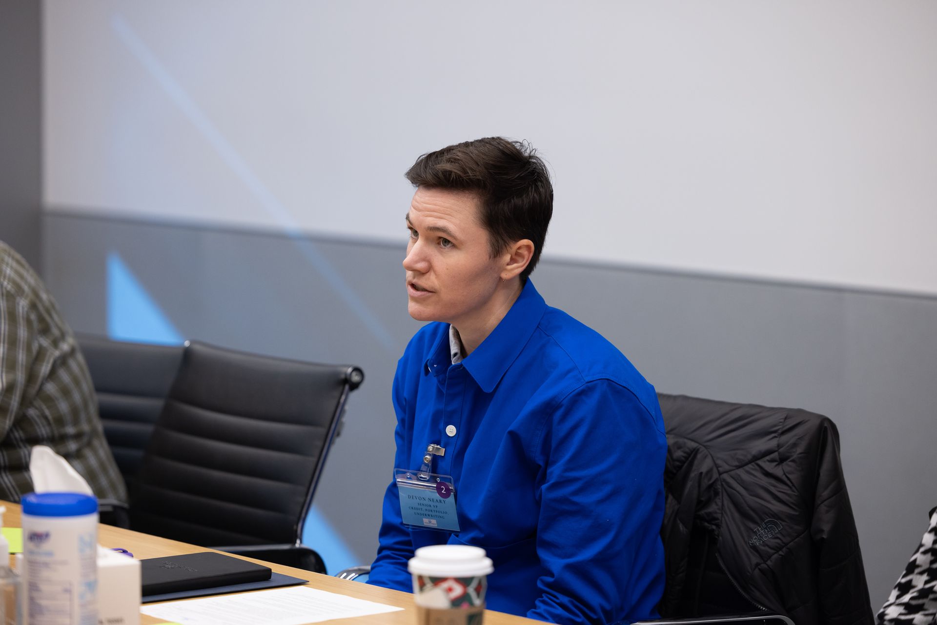 Person in a blue shirt seated at a conference table in a meeting room, listening attentively.