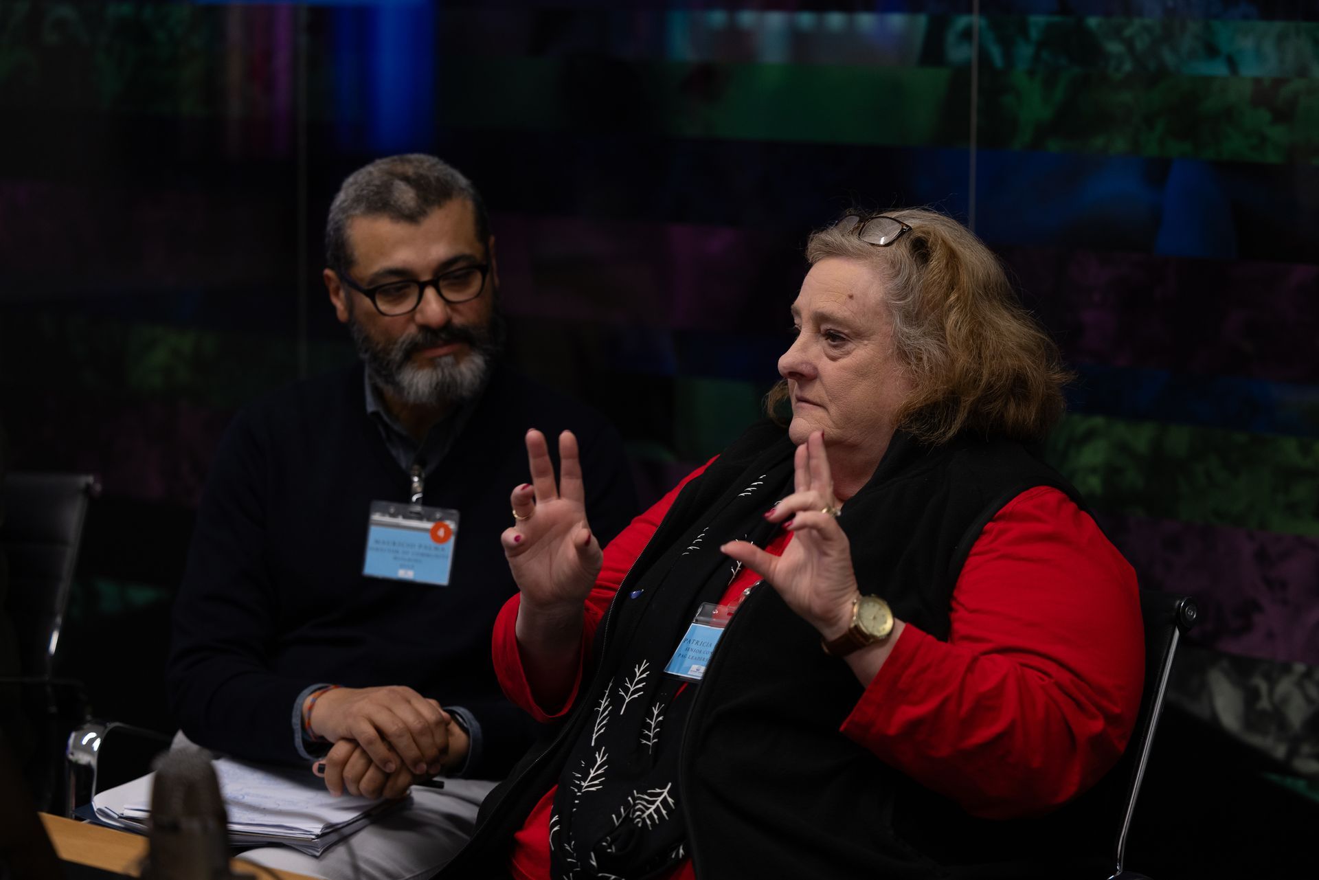 Two people seated at a panel, one speaking with raised hands in a dark conference setting