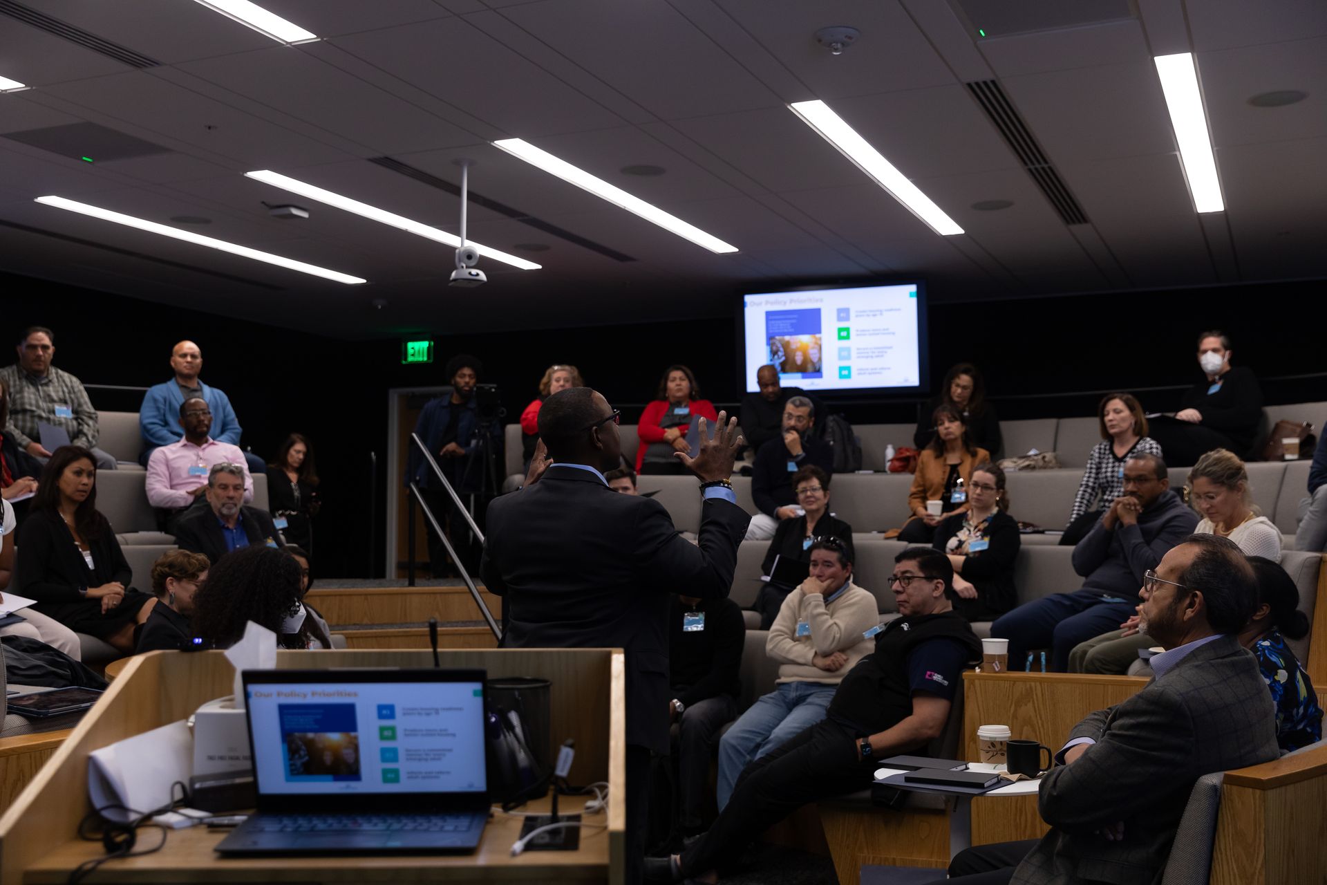 Meeting room with seated audience watching a presenter beside a screen, with a laptop in the foreground