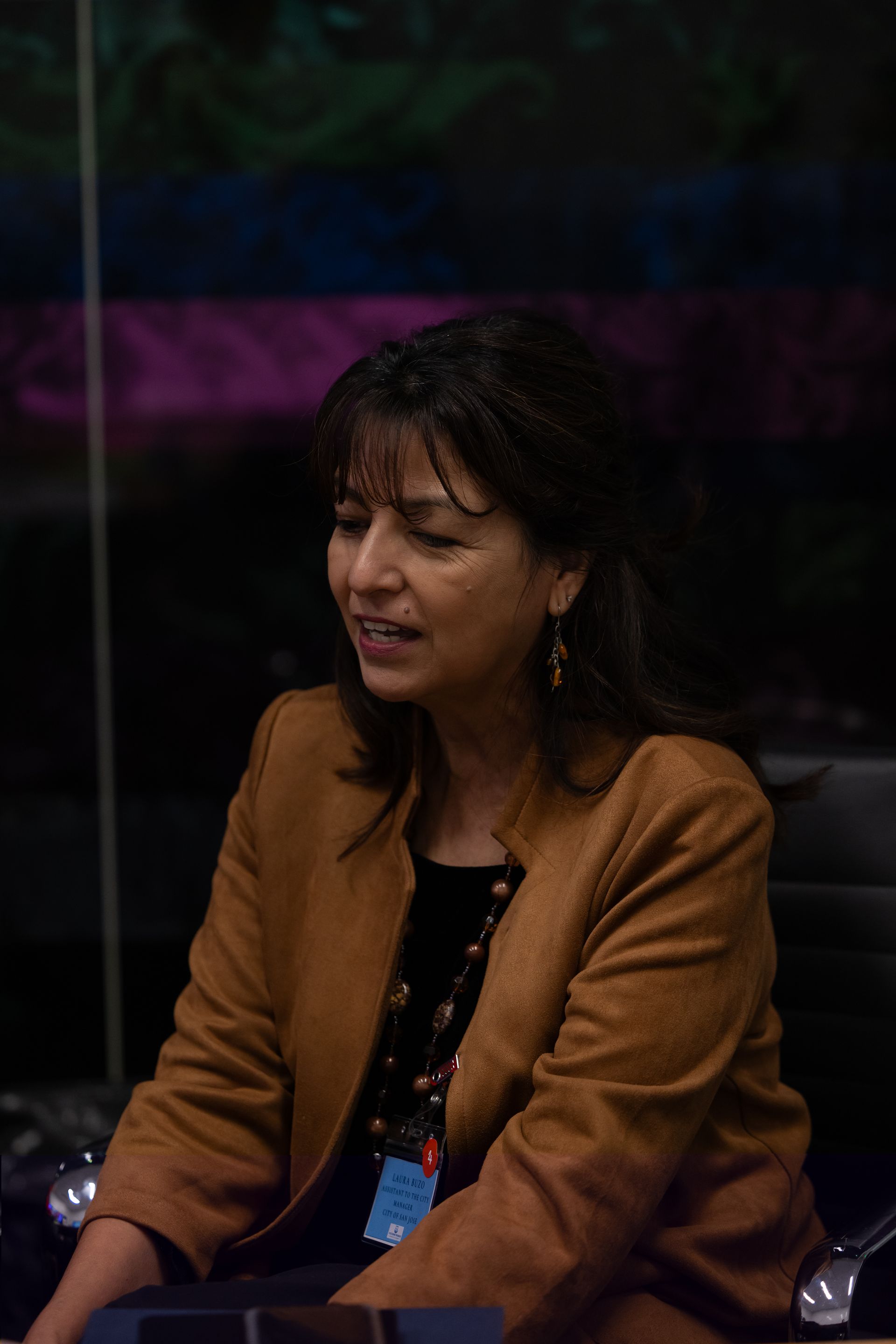 Woman in a brown blazer seated onstage, speaking into a microphone against a dark background.