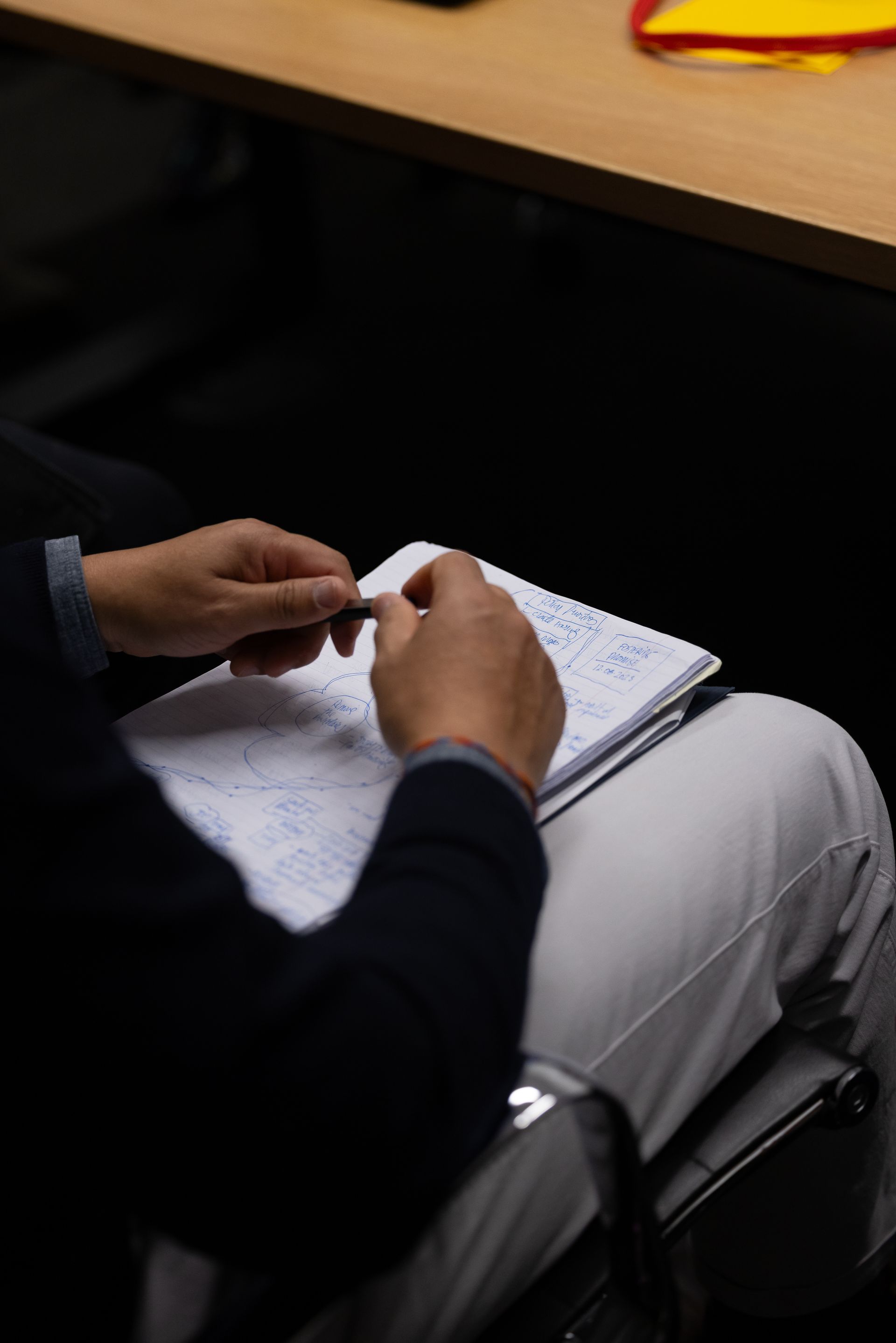 Person writing in a notebook at a desk, with hands visible and a black chair nearby.