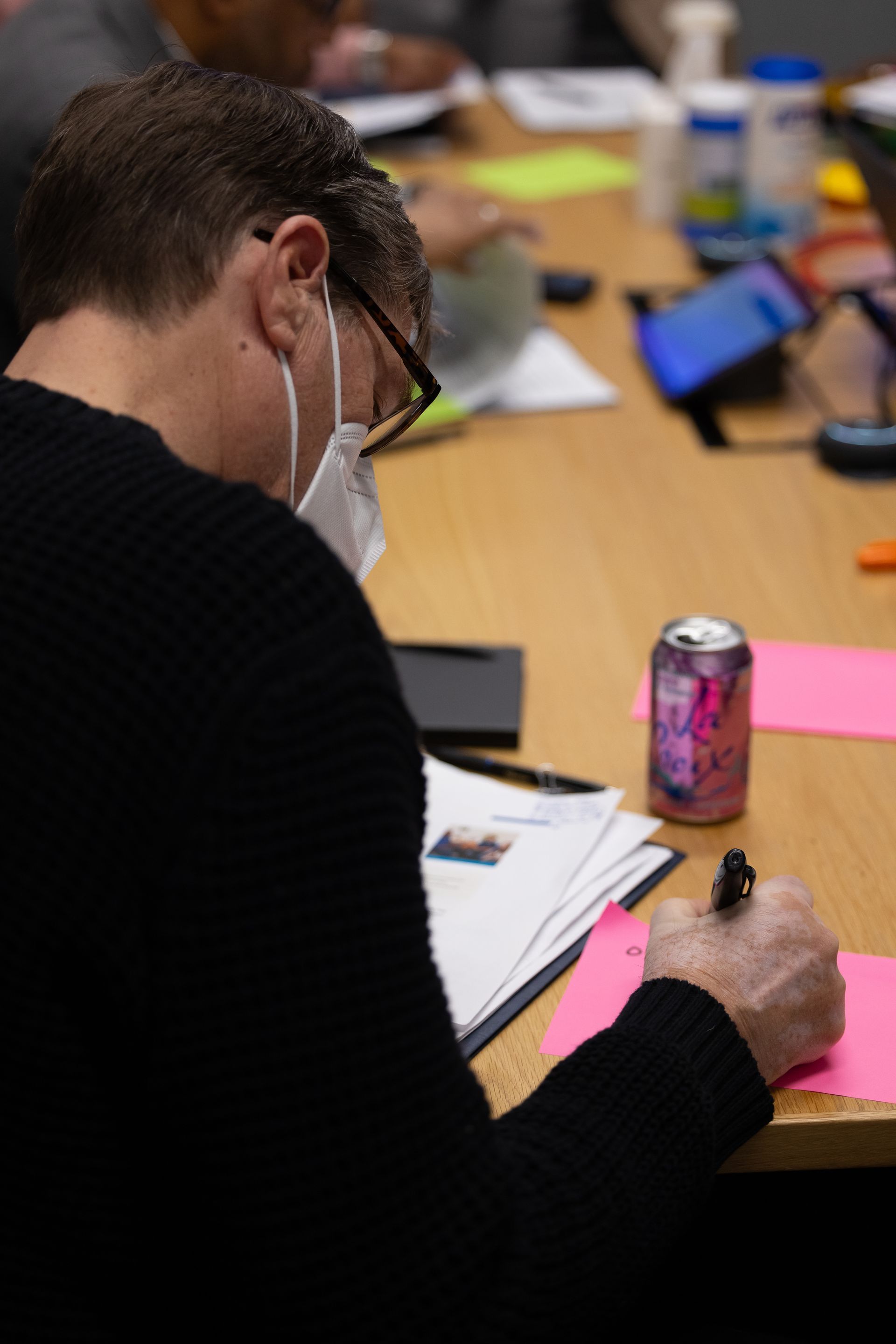 Person wearing a mask writes at a table with papers, a pink note, and a soda can.