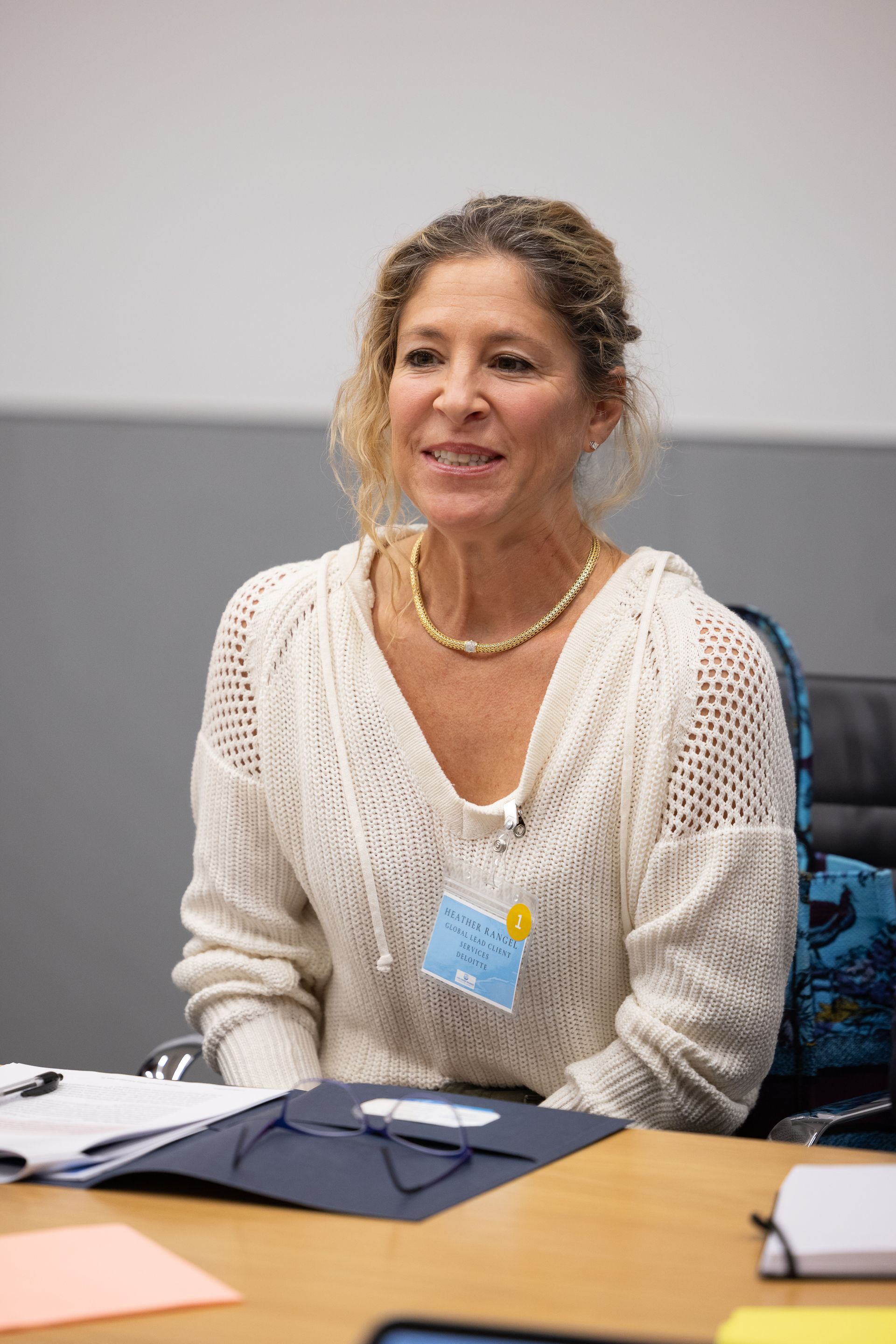 Woman seated at a conference table, wearing a white sweater and lanyard, smiling attentively.