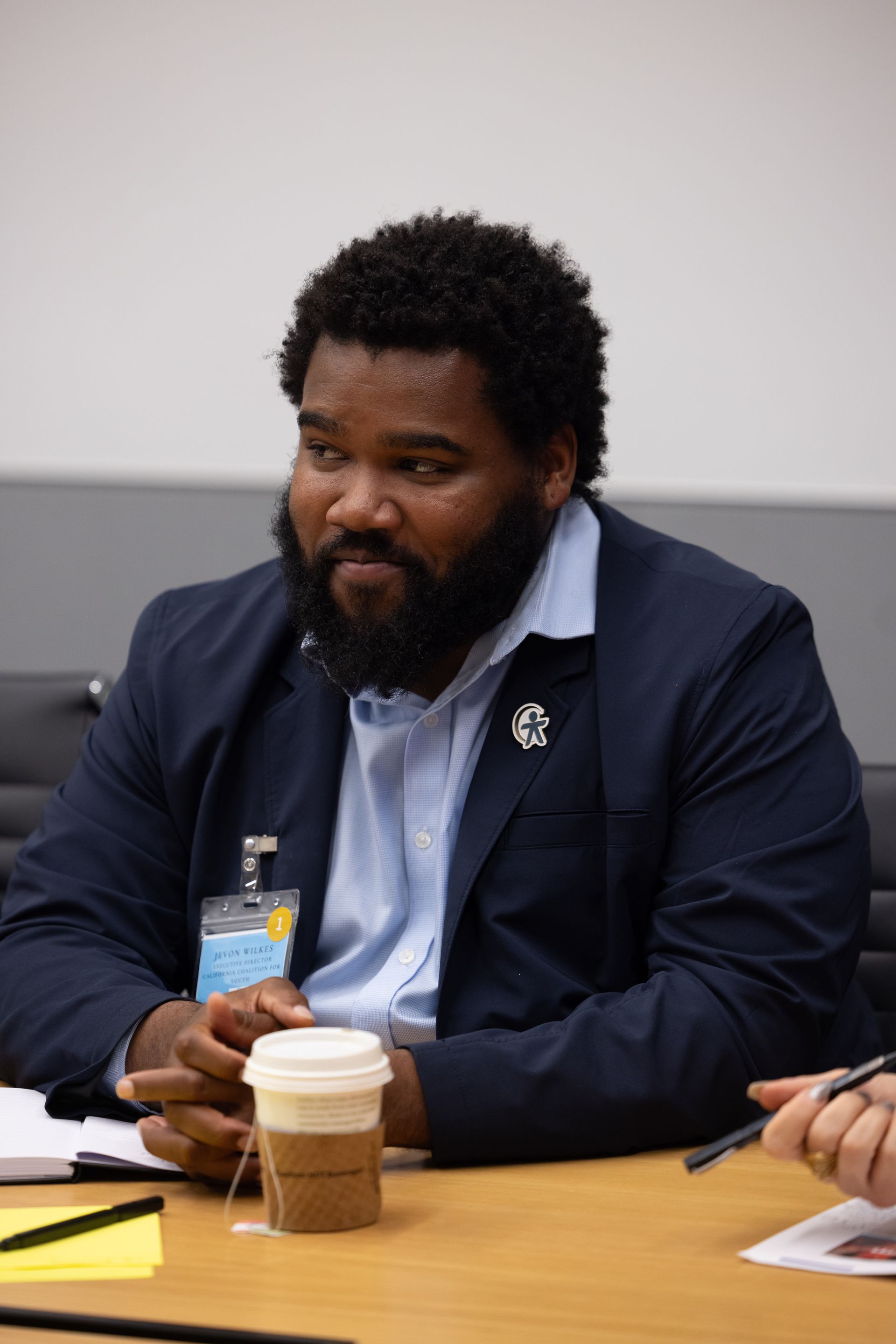 Man in a navy blazer sits at a conference table with a coffee cup and notes.