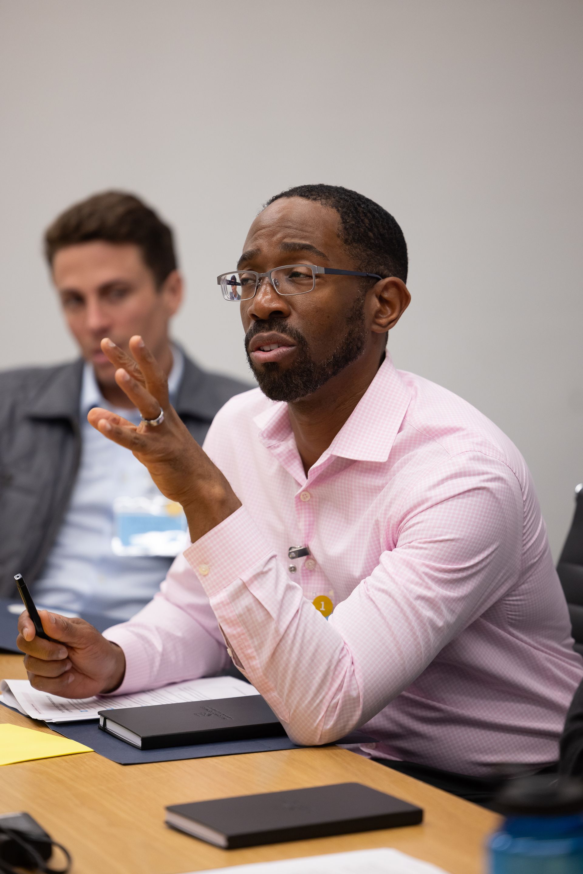 Man in pink shirt speaking at a meeting table, holding a pen and gesturing while others listen nearby