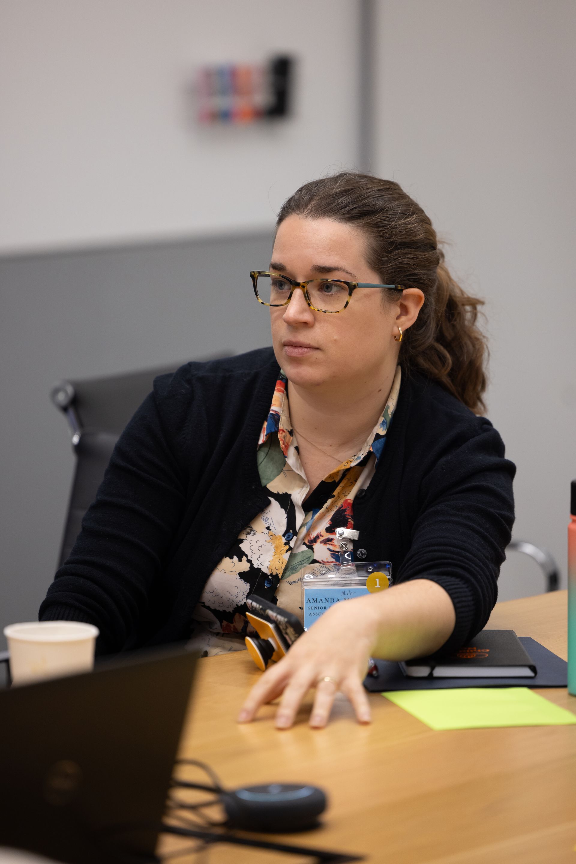 Woman seated at a conference table, looking left and gesturing with her hand in a meeting room.