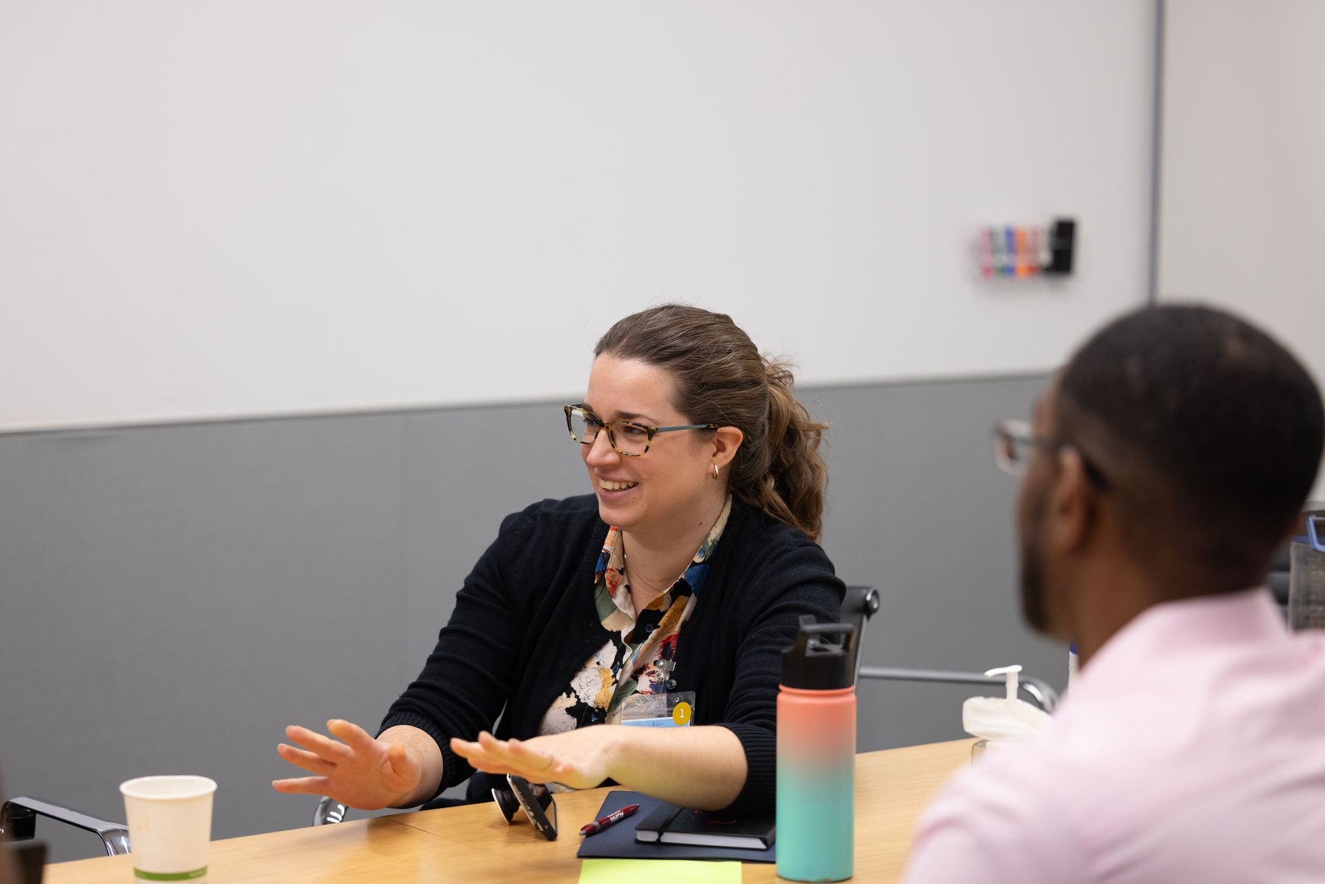 Two people talking at a table in a meeting room, with cups and a colorful water bottle.