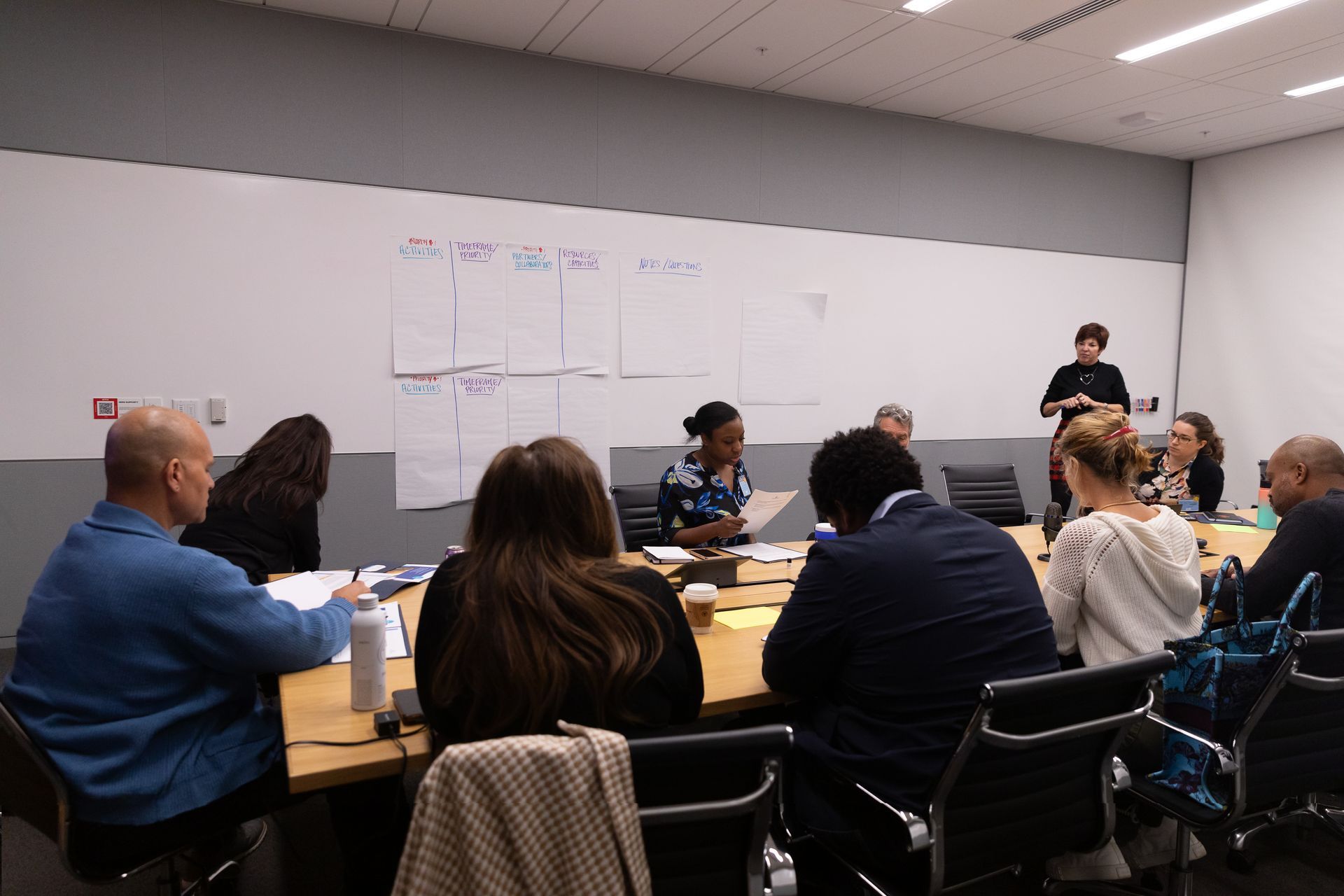 People seated at tables in a conference room watching a speaker near a whiteboard.