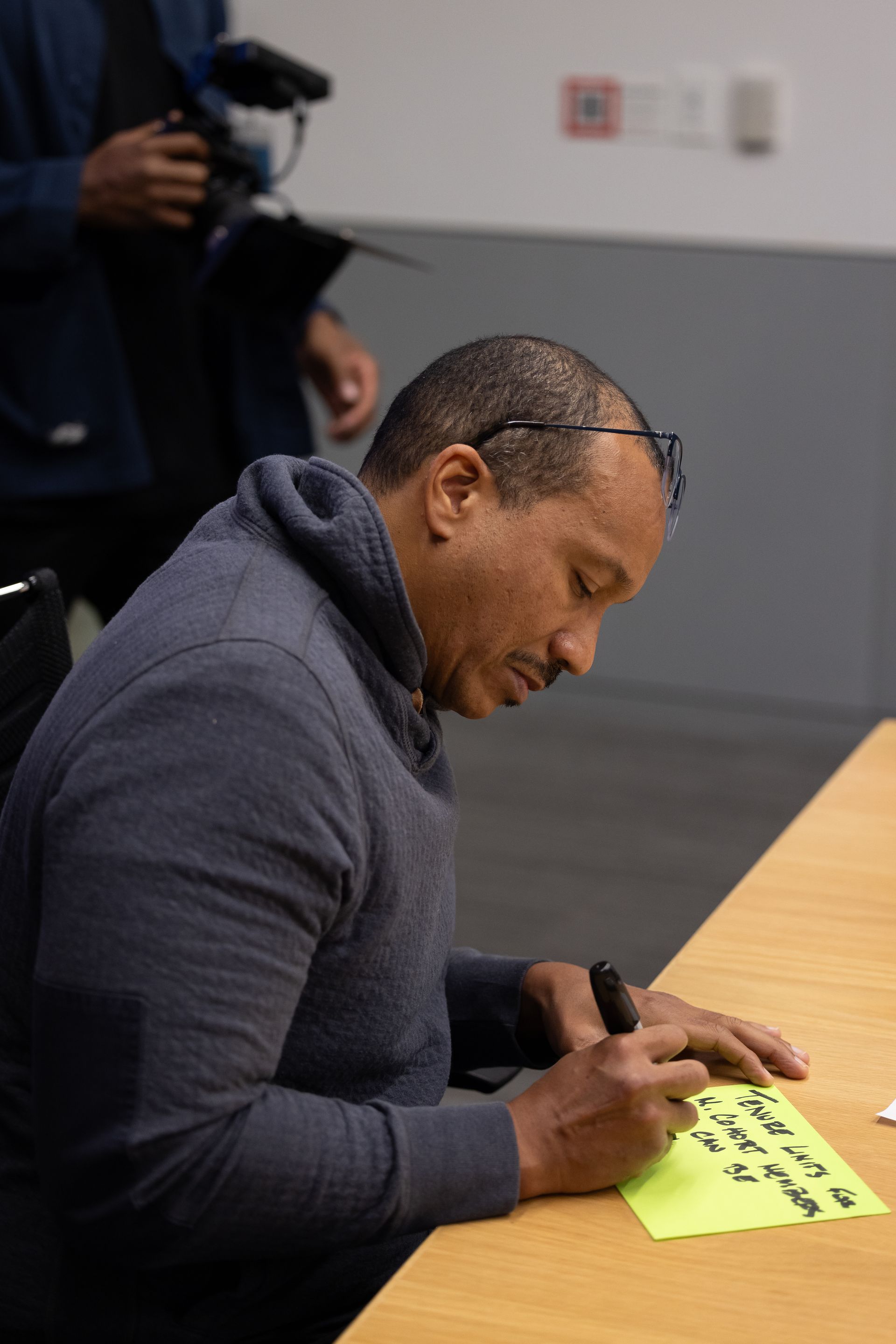 Man signing a yellow sheet at a table, with a photographer in the background.