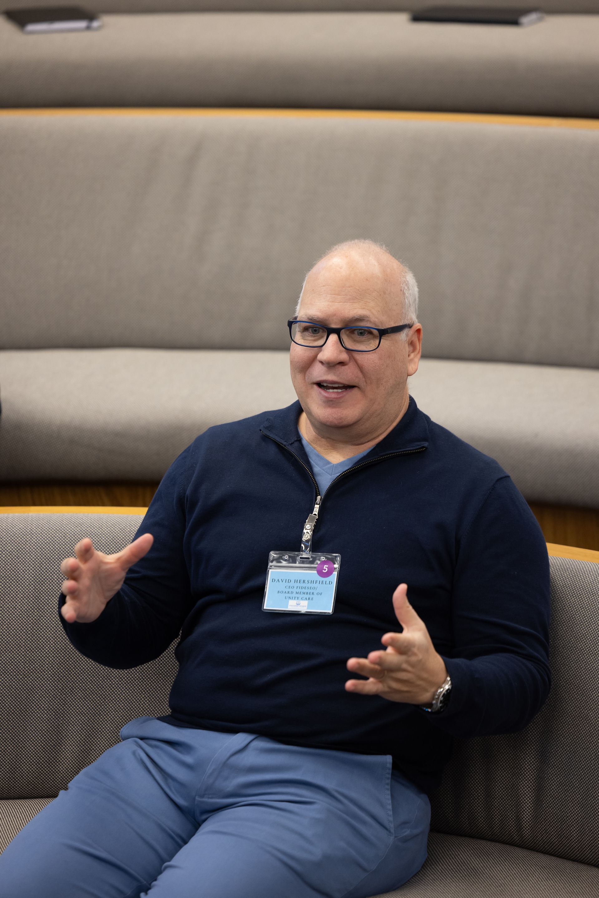 Man speaking and gesturing while seated on a couch in a modern lounge area.