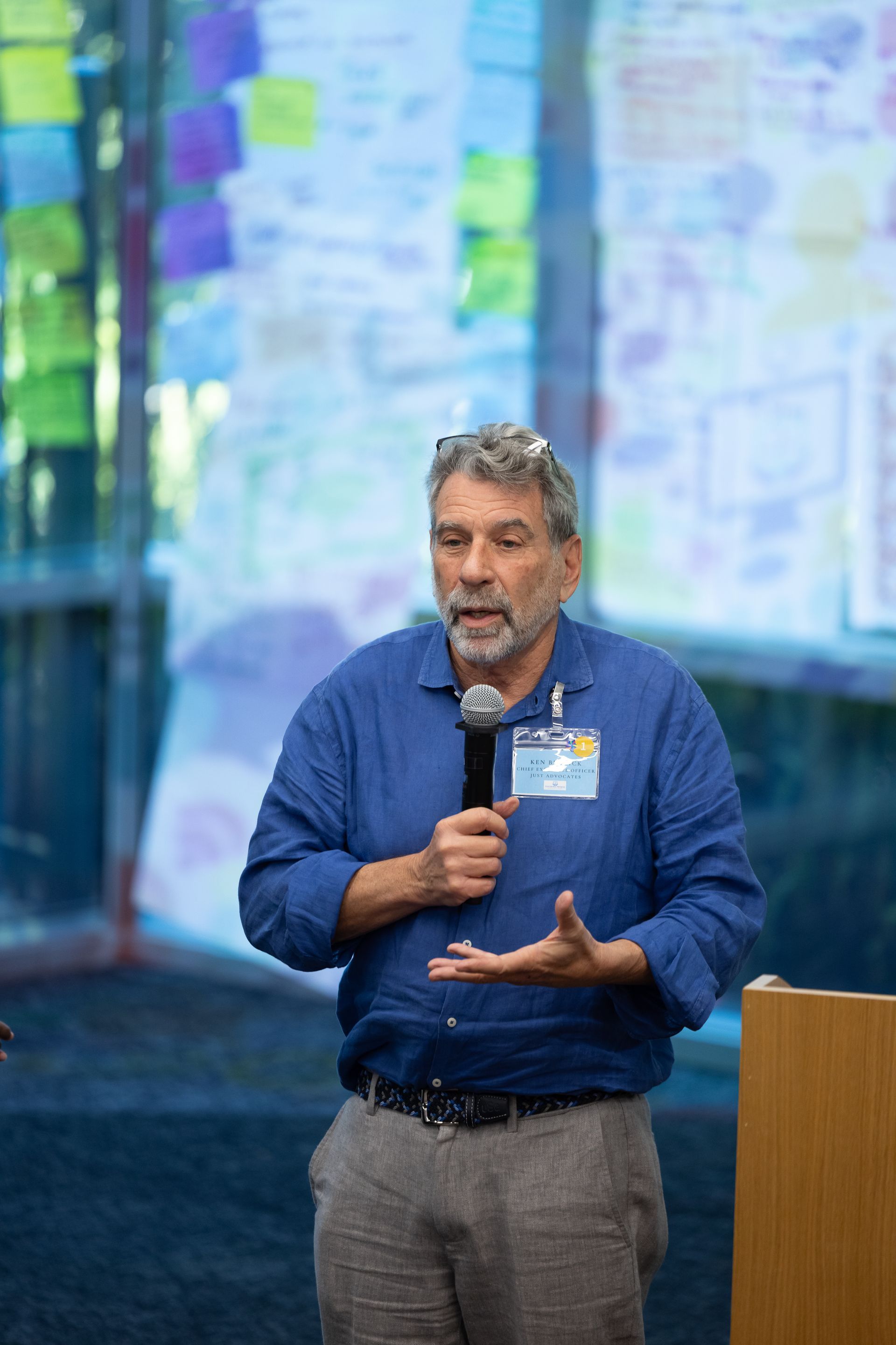 Man in blue shirt speaking into a microphone at a conference podium with presentation screens behind him