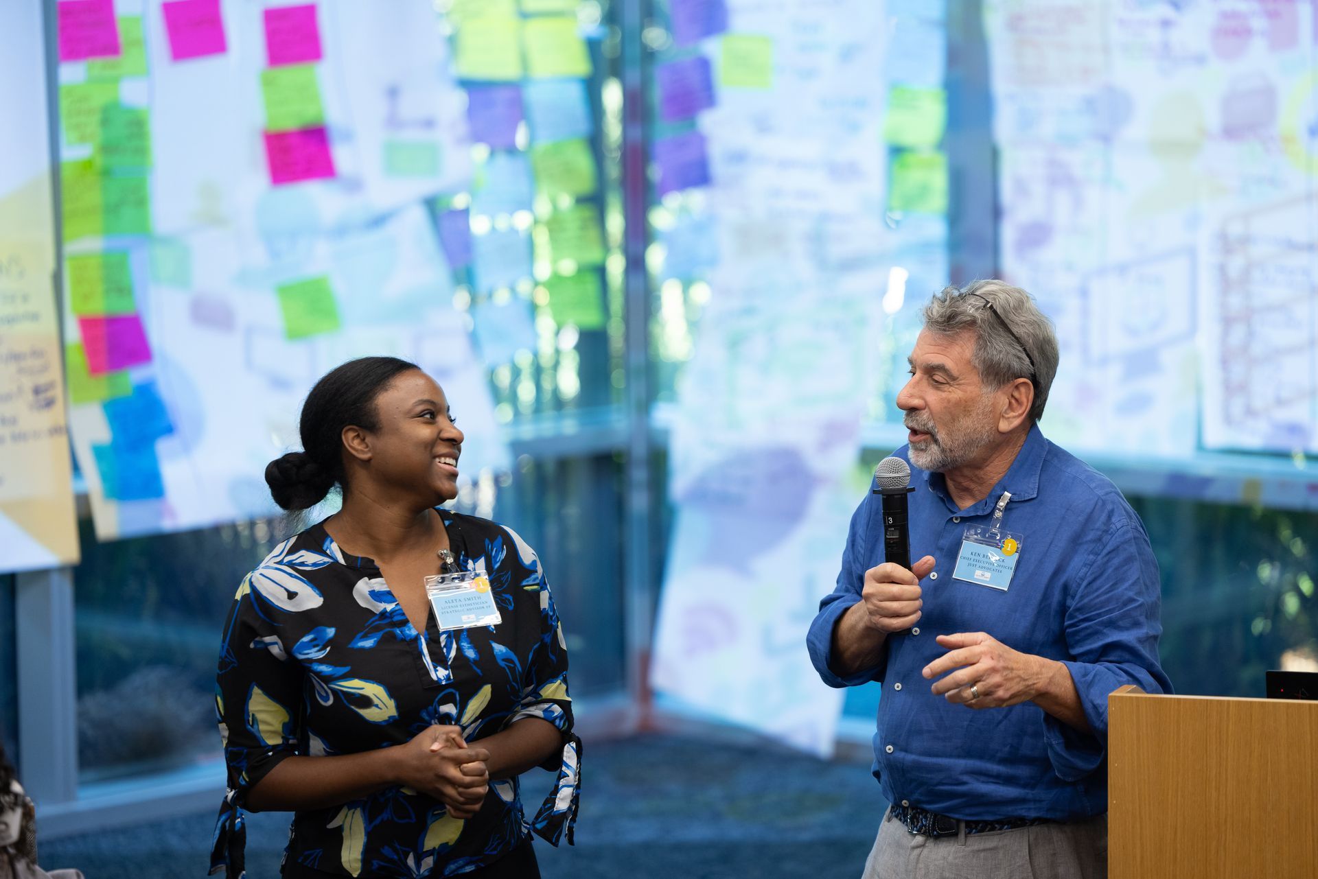 Two speakers converse on a stage in front of colorful sticky notes and a presentation screen.