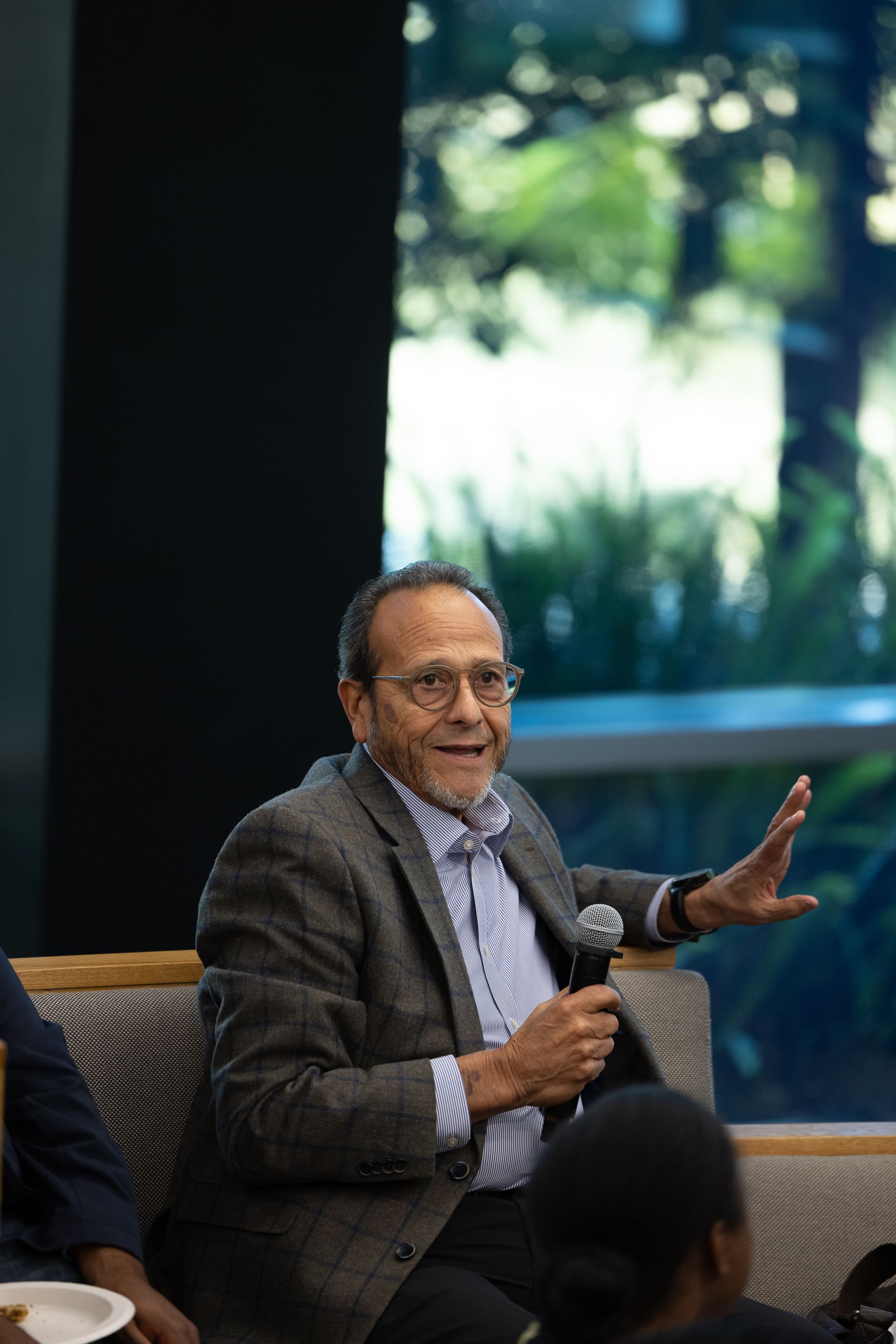 Man speaking into a microphone while seated on a chair in a bright indoor setting