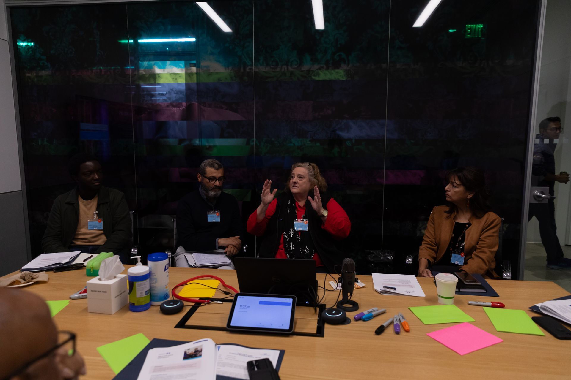 People seated at a conference table; a woman in a red jacket speaks while others listen in a meeting room.
