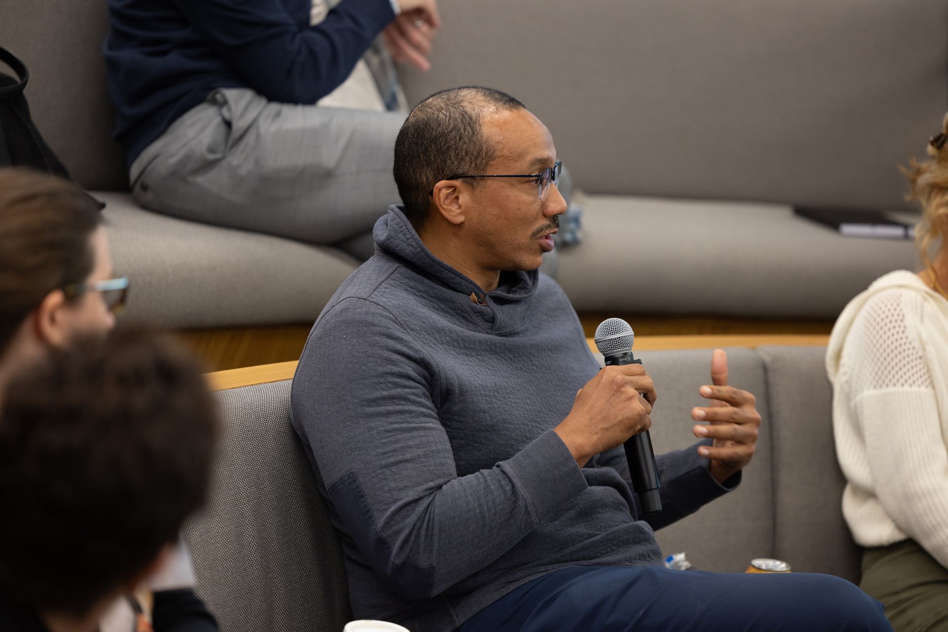 Man in a gray sweater speaking into a microphone during a seated discussion