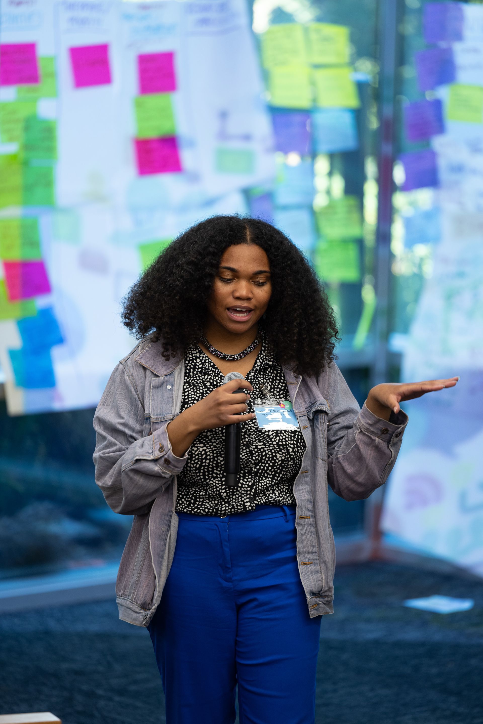 Woman speaking on stage with a microphone, gesturing in front of colorful sticky notes on a glass wall.