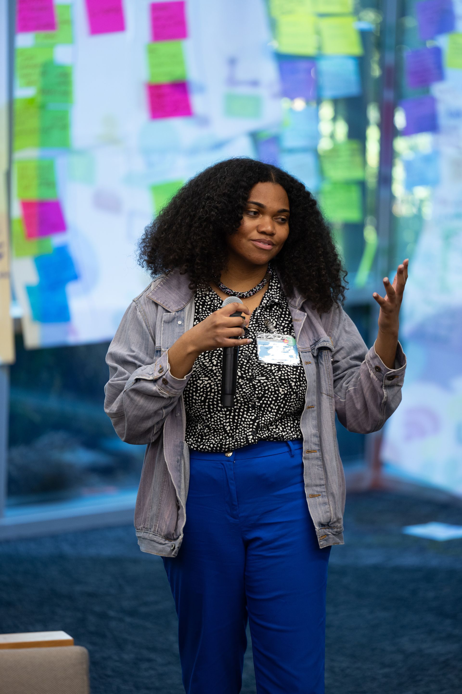 Woman speaking into a microphone at an indoor event, wearing a striped jacket and bright blue pants.