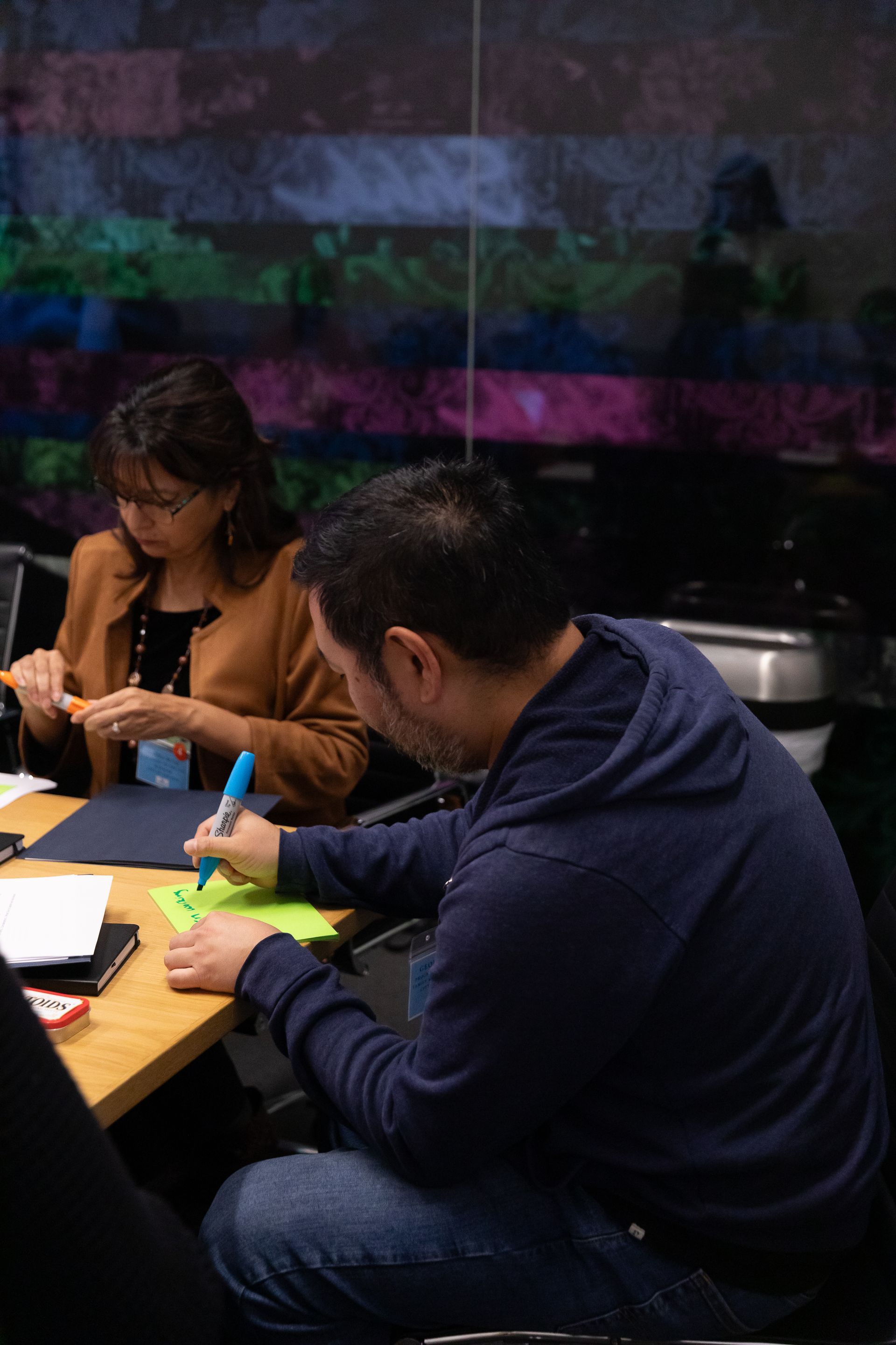 Two people seated at a table in a dim cafe, writing and reading notes beside drinks.