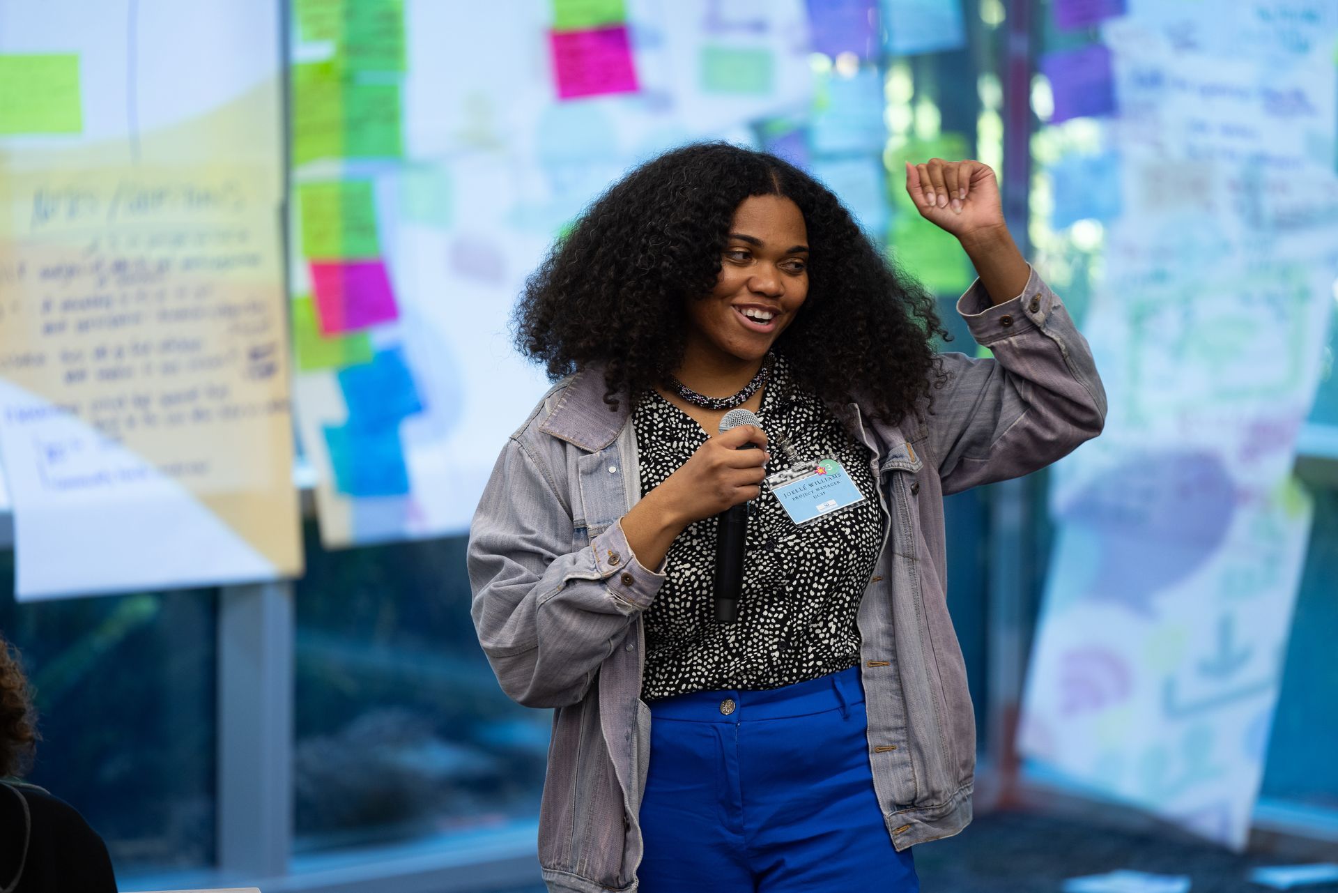Woman speaking with a microphone and raising her fist in front of a wall covered with colorful notes.