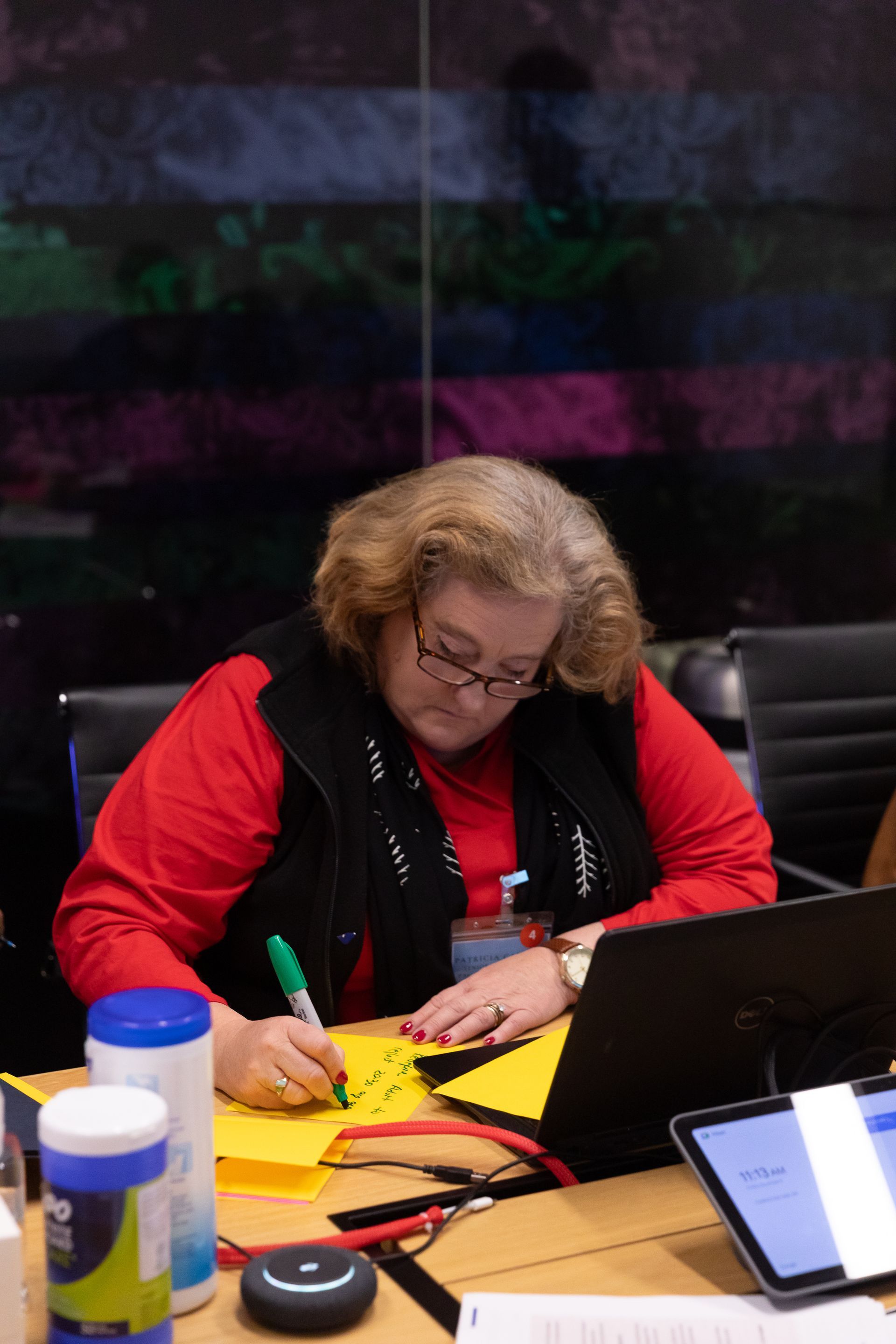 Person writing at a conference table with a laptop, papers, and a coffee cup nearby.