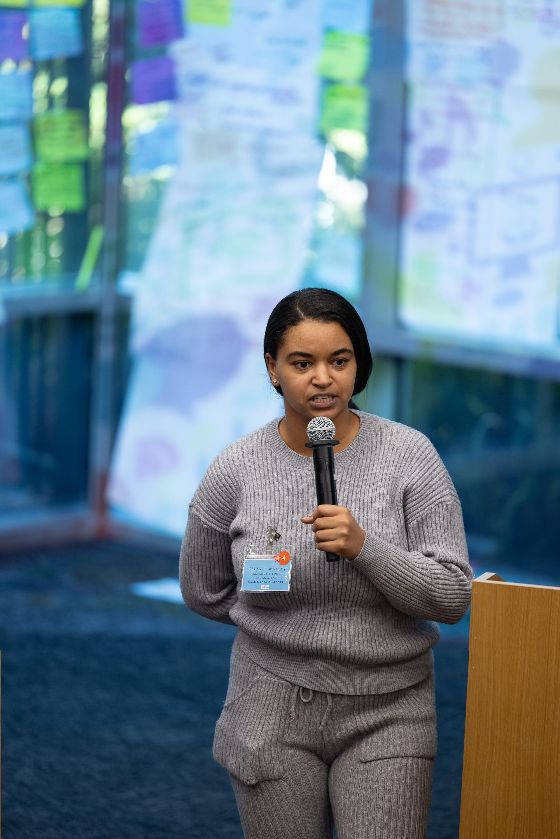 Woman speaking into a microphone on a stage, wearing a gray sweater and matching pants, with a blurred blue background.