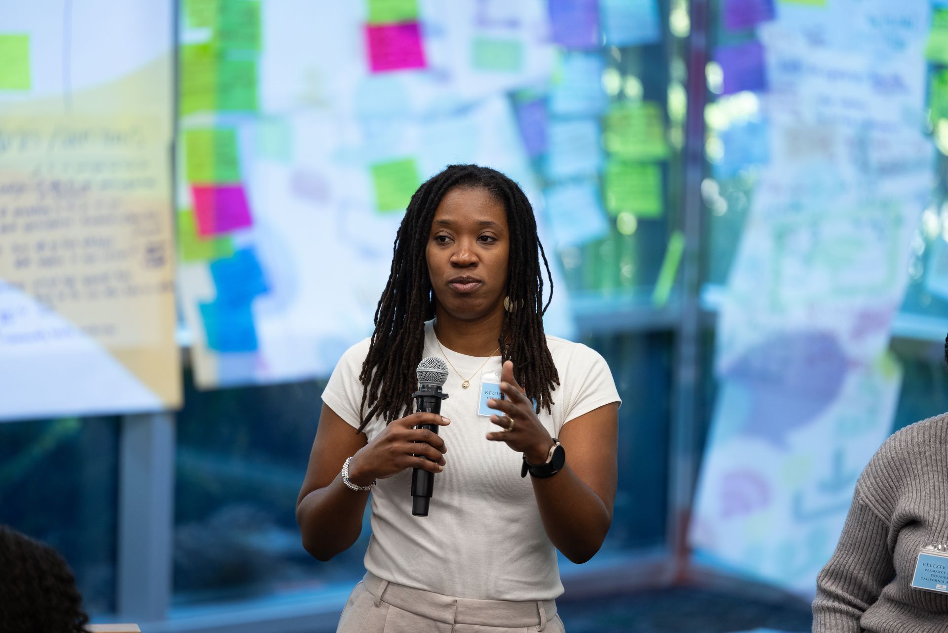 Woman speaking into a microphone at an event, with colorful notes and charts in the background.