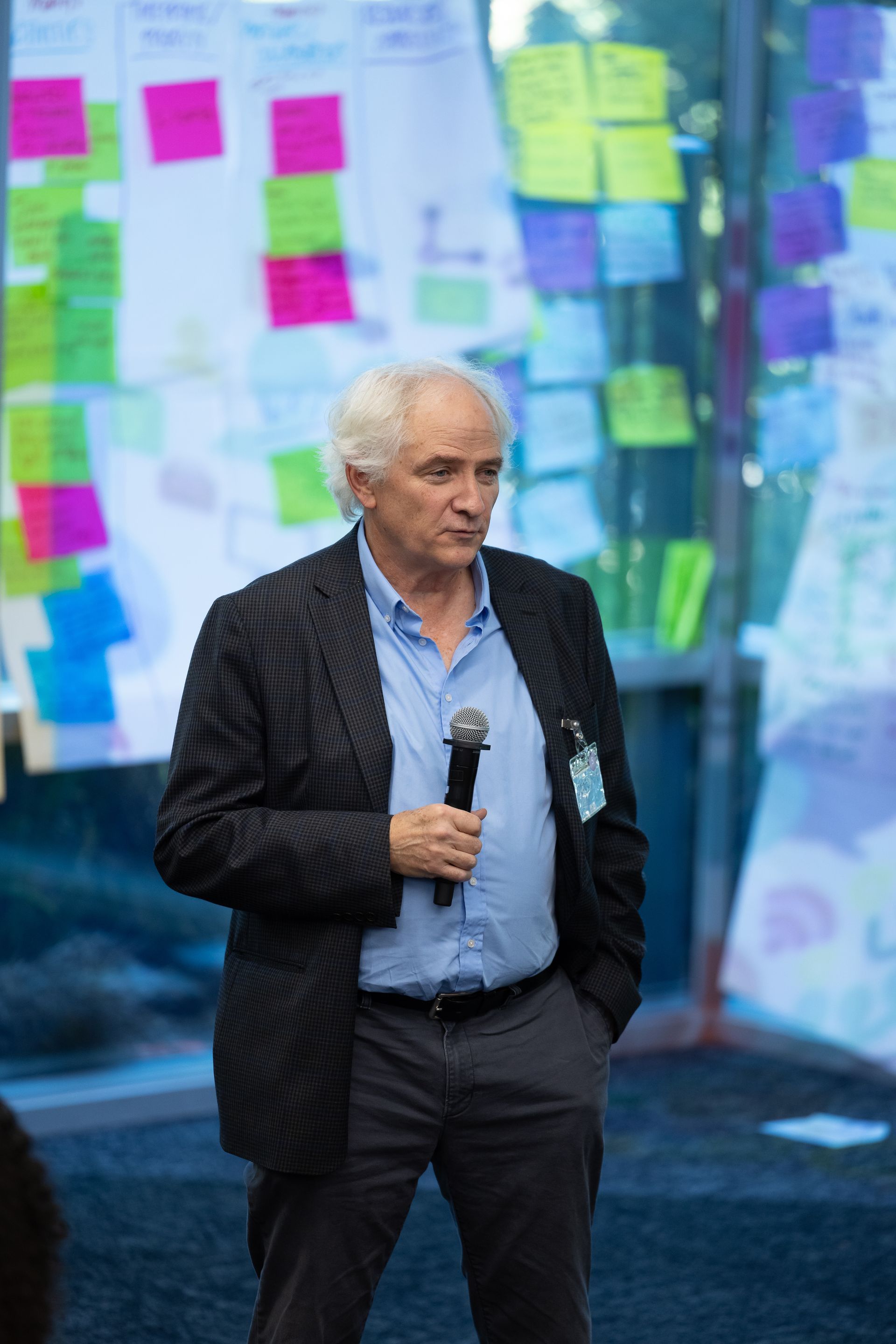 Man standing onstage holding a microphone in front of a colorful wall of sticky notes