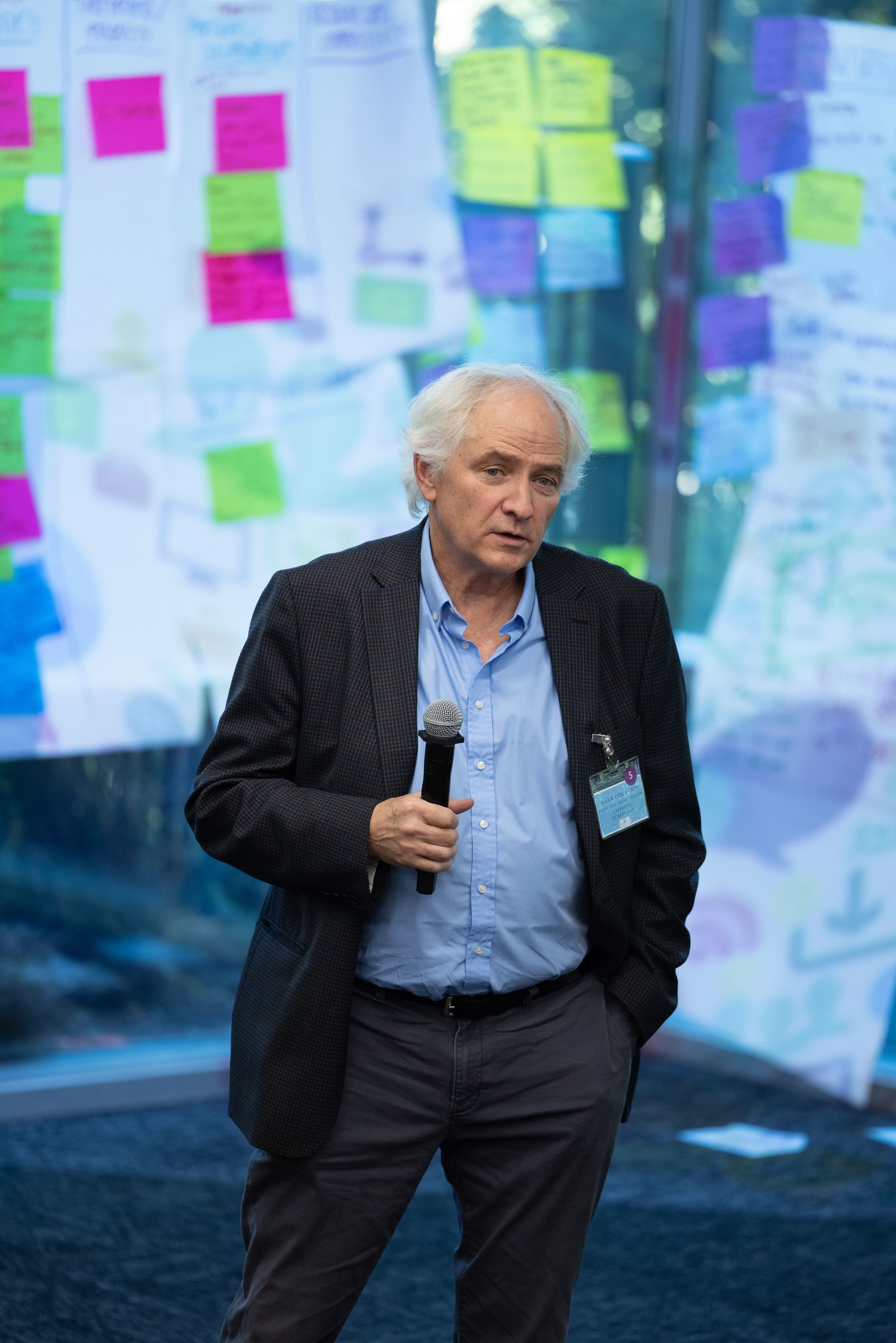 Man speaking onstage with a microphone, in front of a colorful wall of sticky notes