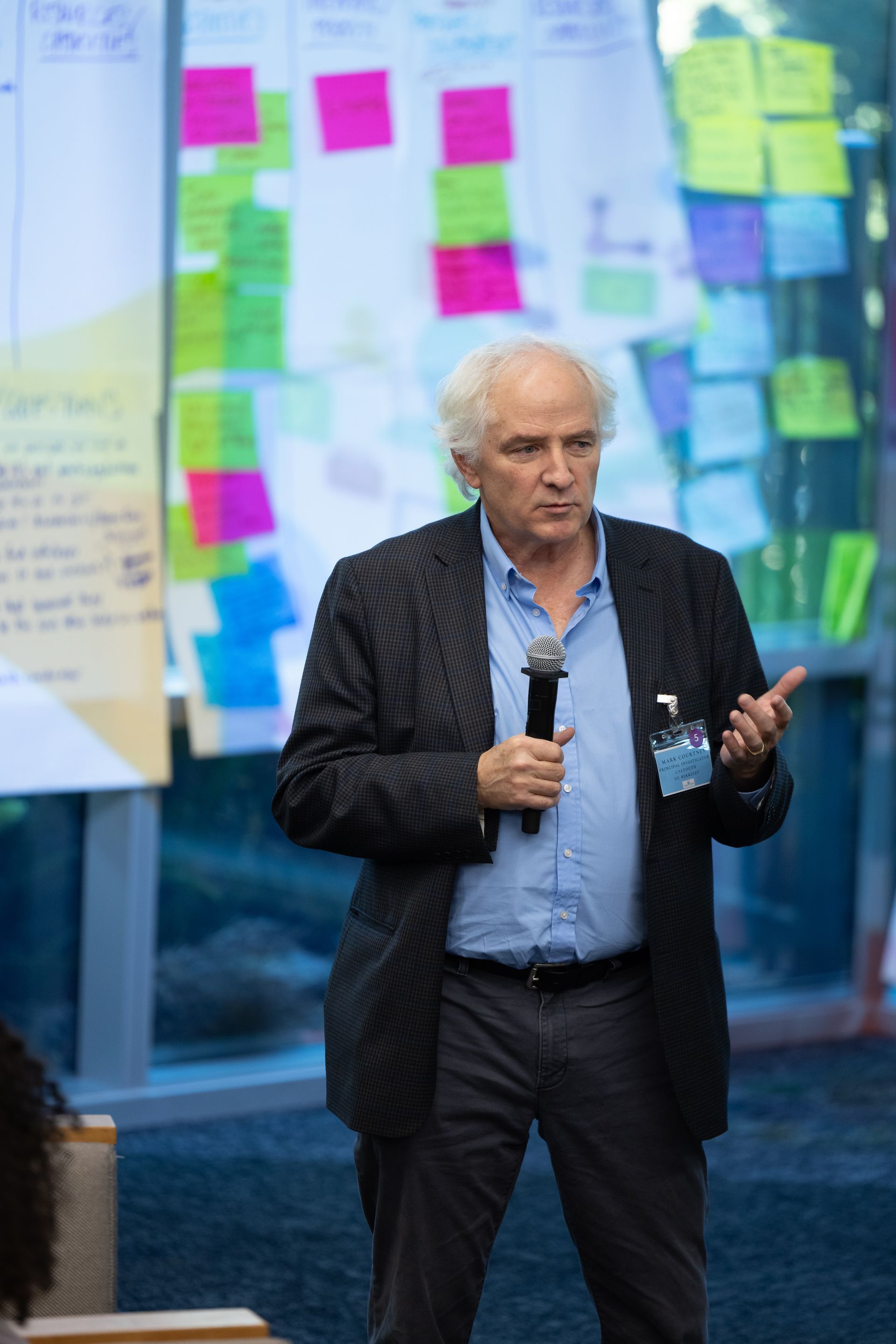 Man speaking into a microphone at a conference, with colorful sticky notes on glass walls behind him