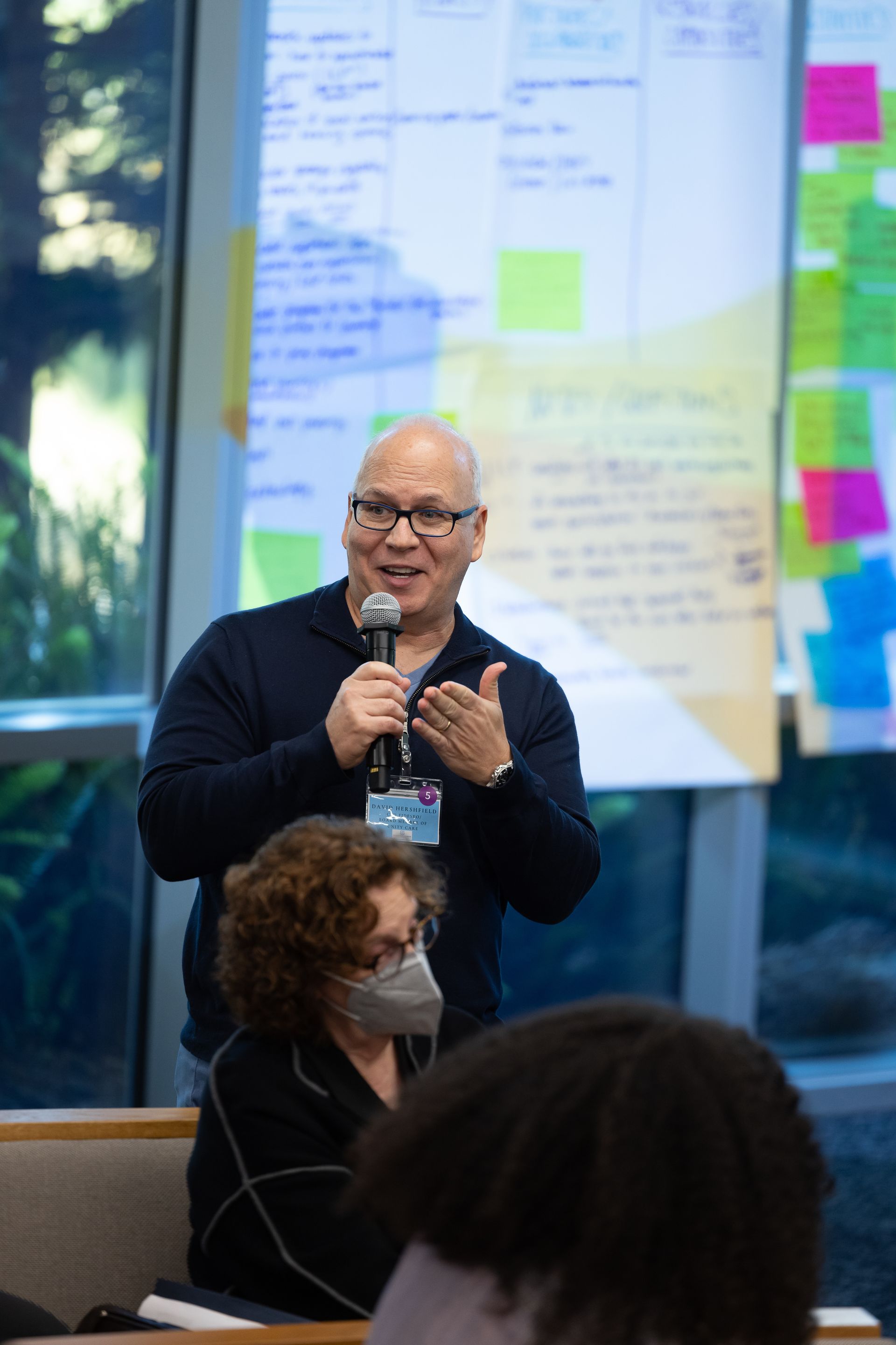 Speaker holding a microphone at an indoor event, with a blurred presentation board and colorful sticky notes behind them