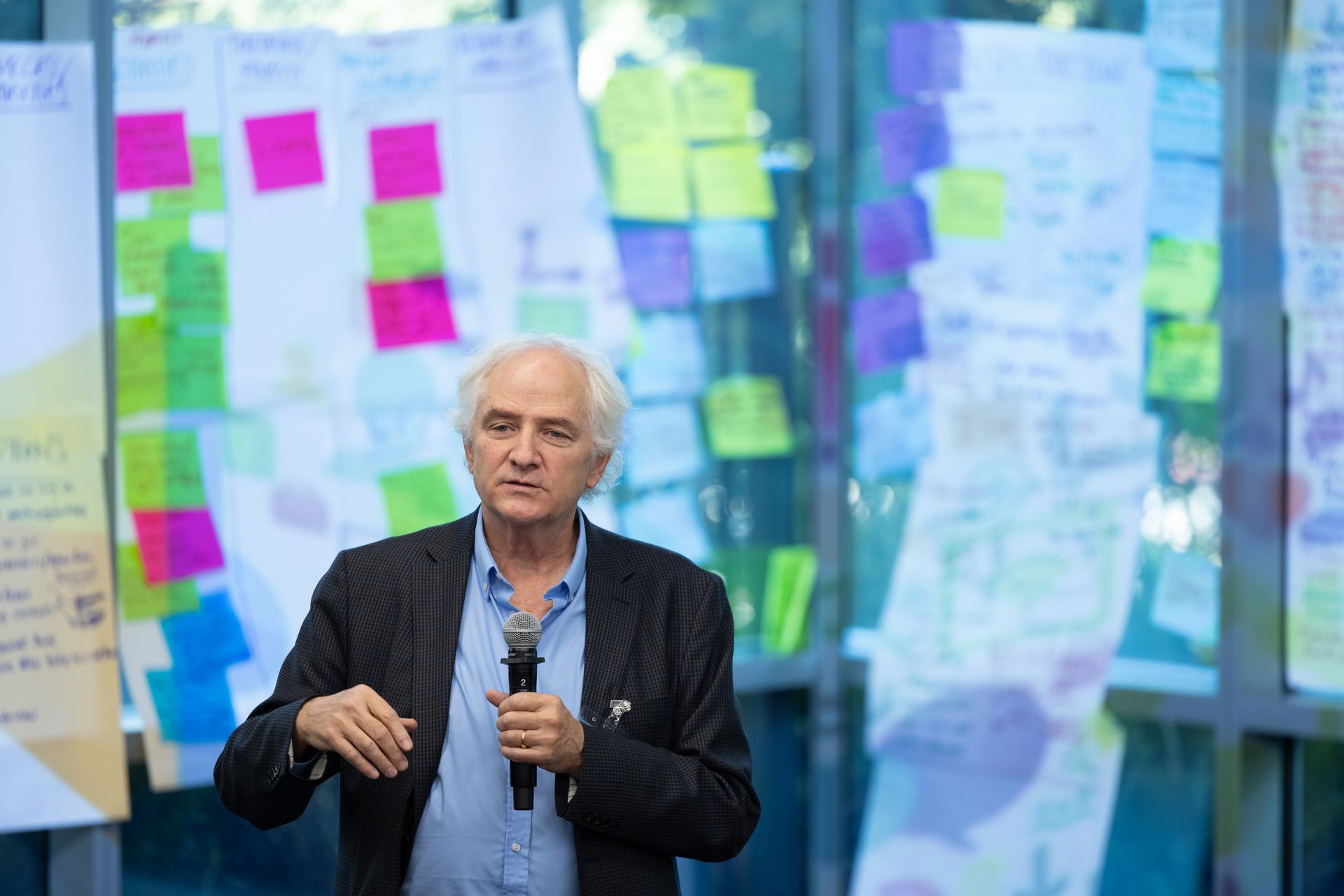 Man speaking onstage in front of colorful sticky notes and blurred presentation boards