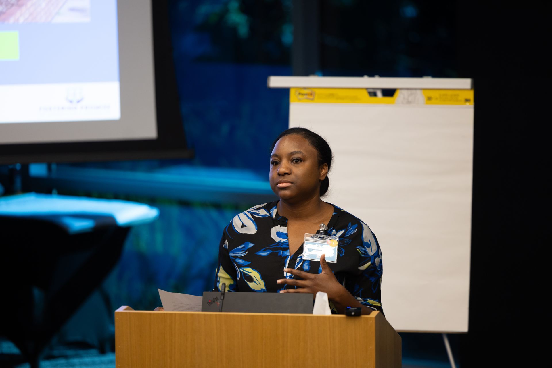 Speaker at podium giving a presentation beside a flip chart and projected screen.