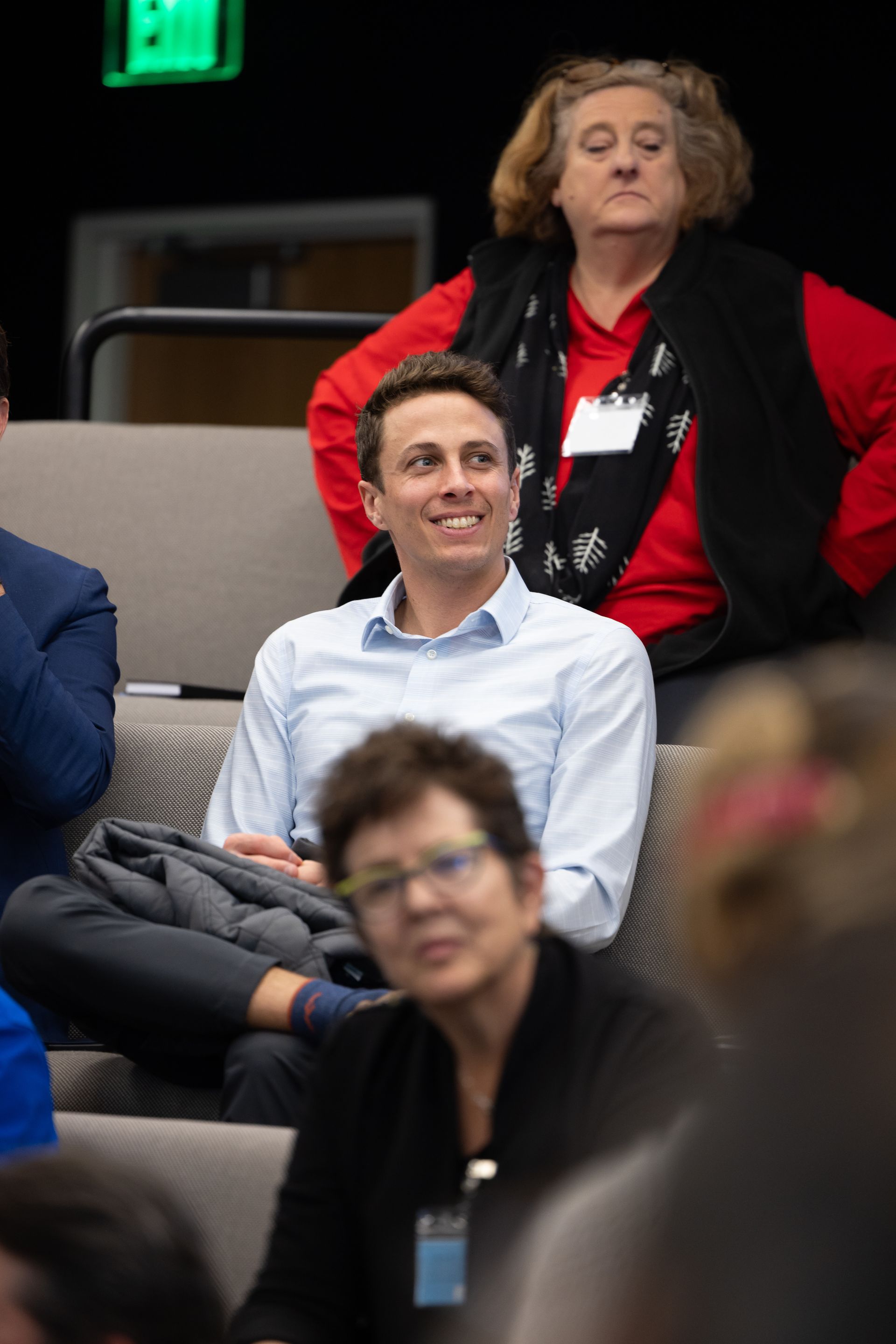 Audience members seated in a lecture hall, one smiling in a light blue shirt with a black jacket on his lap