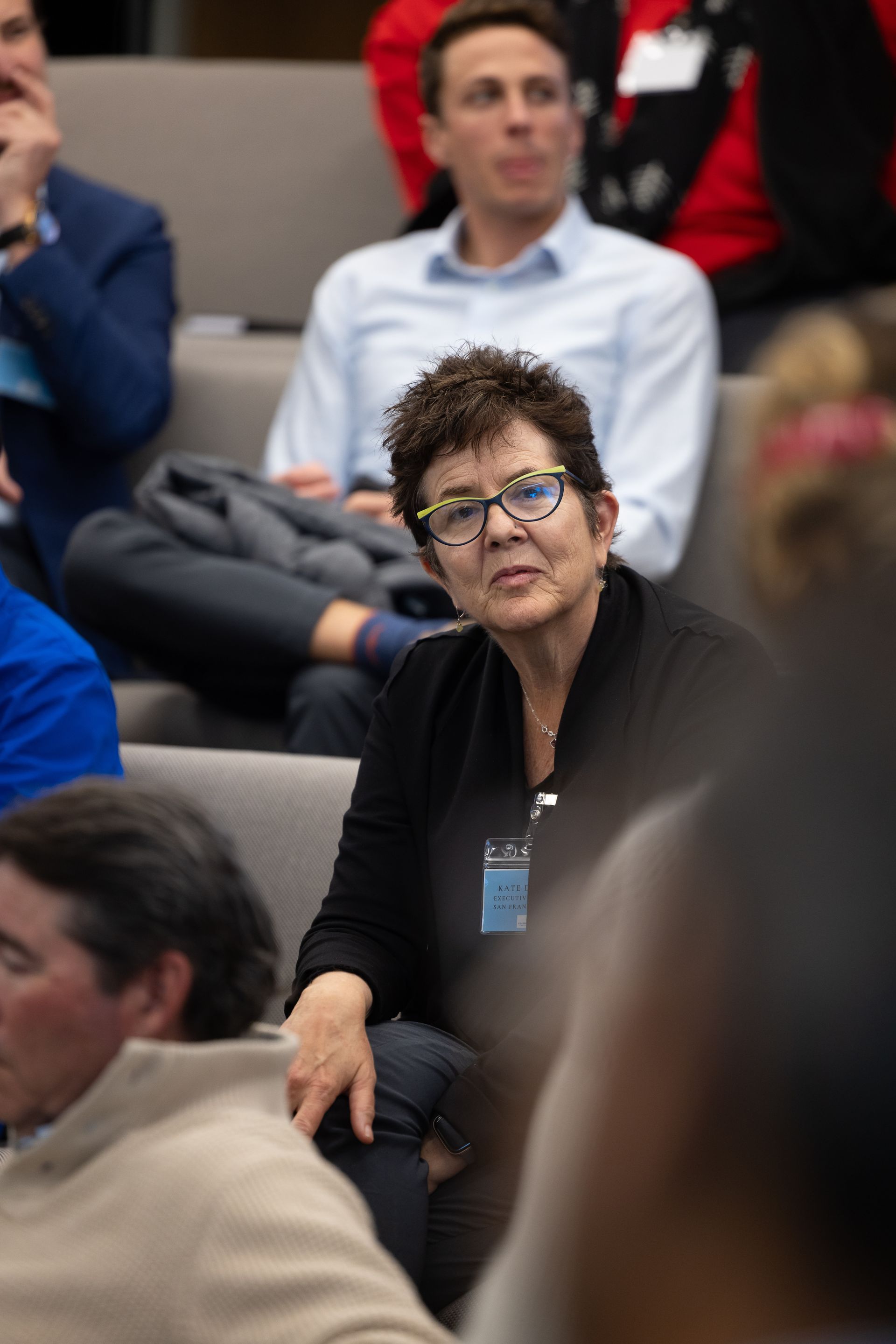 Audience member seated in a crowd, wearing glasses and a black jacket, looking toward the event