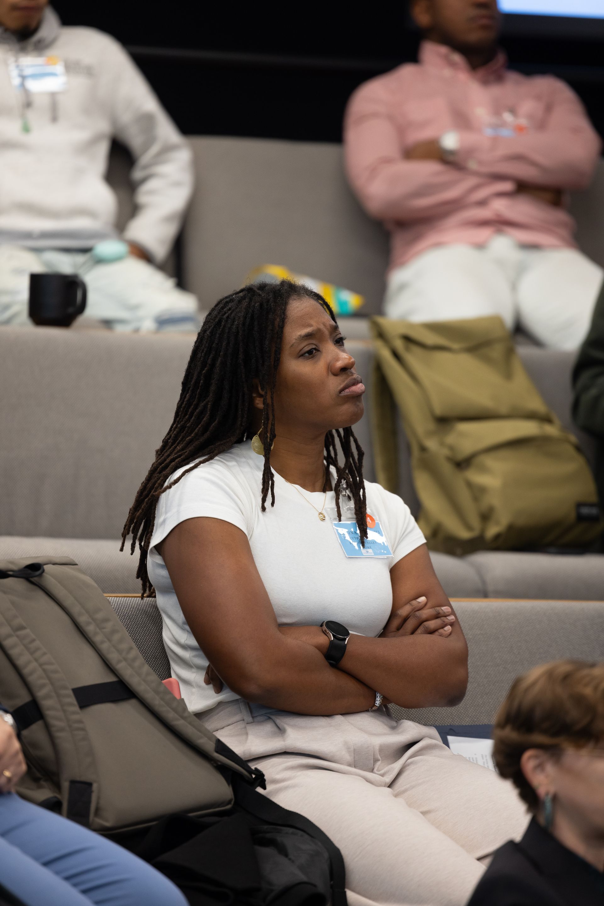 Woman in a white shirt seated with arms crossed at a conference, watching attentively.