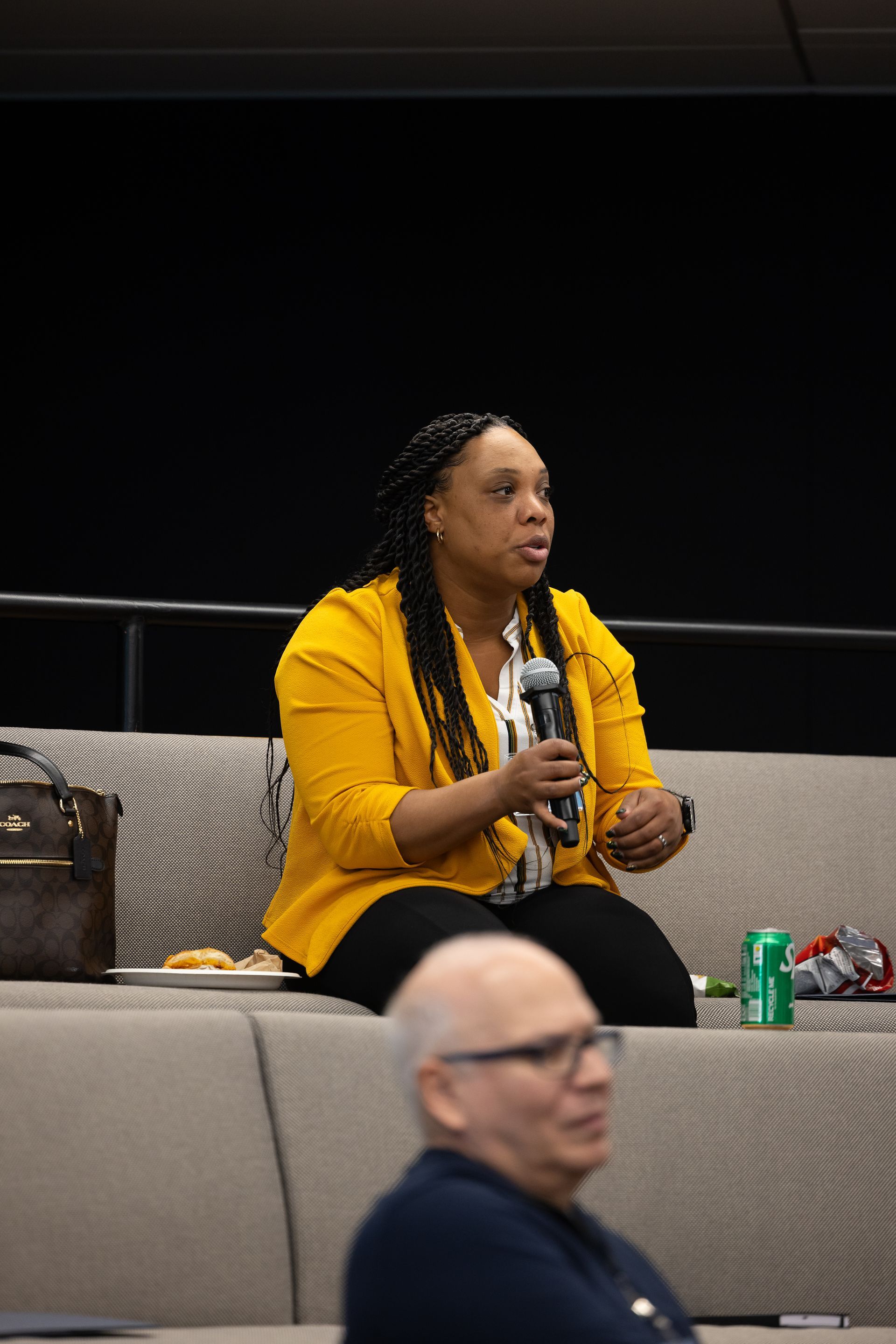 Woman in a yellow blazer speaking into a microphone on a couch in a dark auditorium