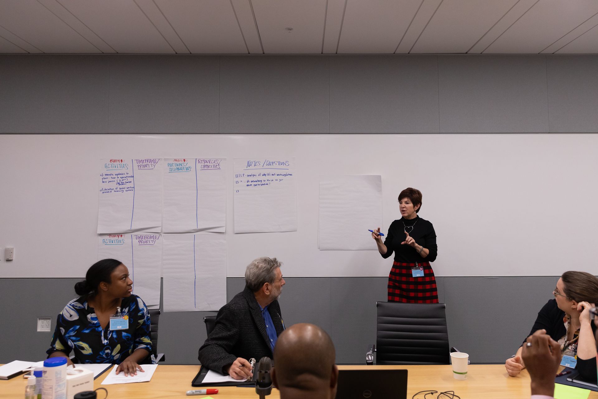 Woman presenting to a small meeting group in a conference room, with charts on the whiteboard behind her.