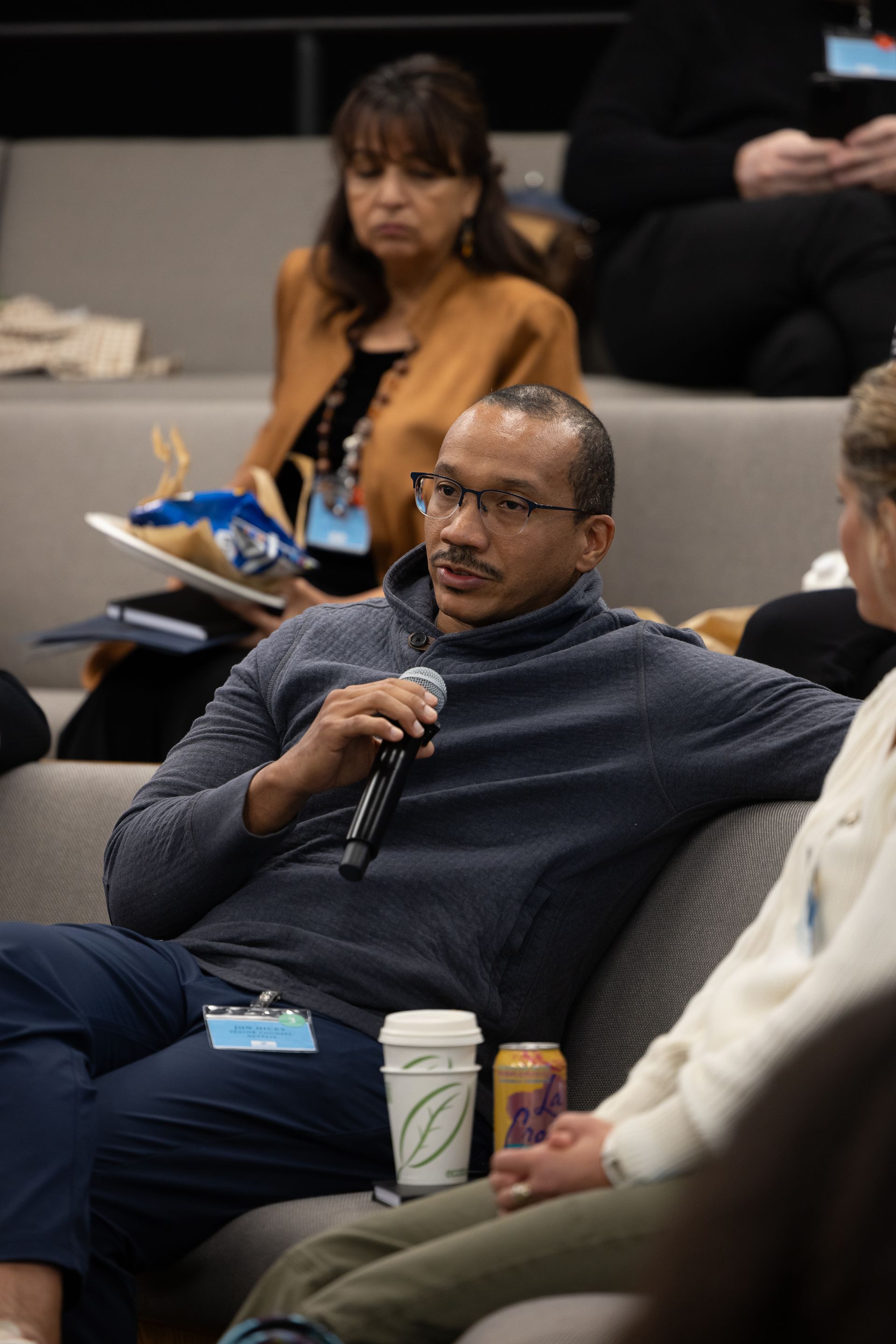 Man speaking into a microphone during a seated discussion, with audience members listening nearby