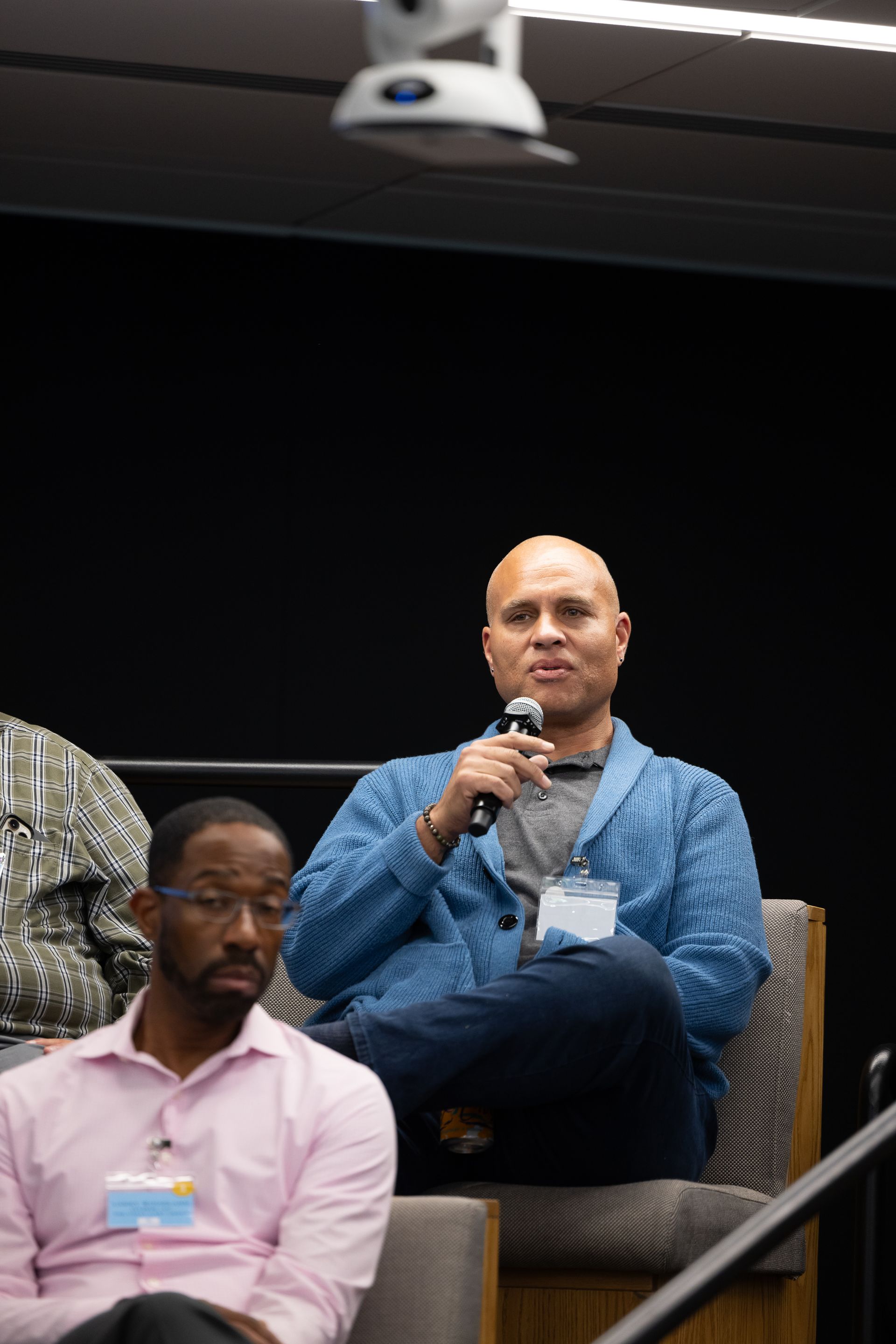 Man holding a microphone while seated onstage in a blue shirt, with others seated nearby.