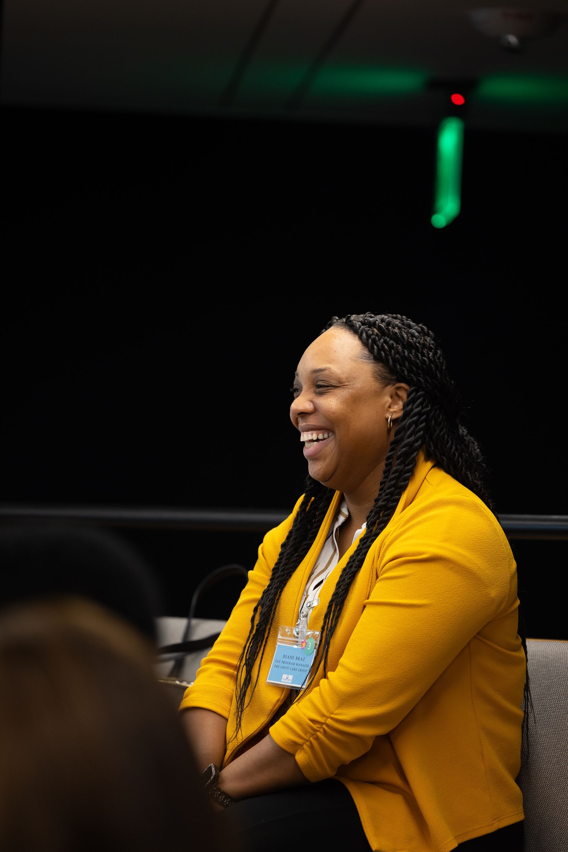 Smiling person in a yellow jacket seated indoors at a dim event or meeting