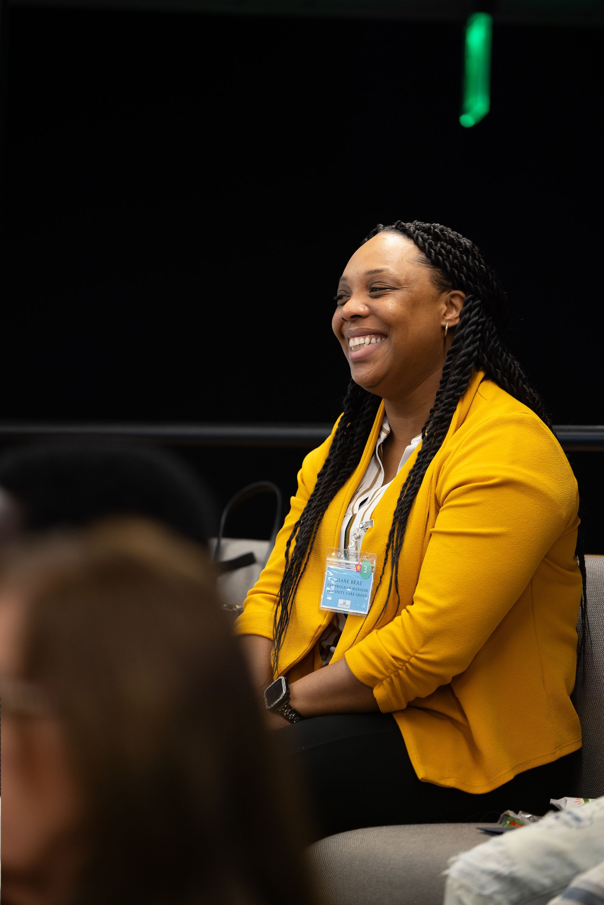 Smiling woman in a yellow blazer seated at an event, looking toward a speaker.