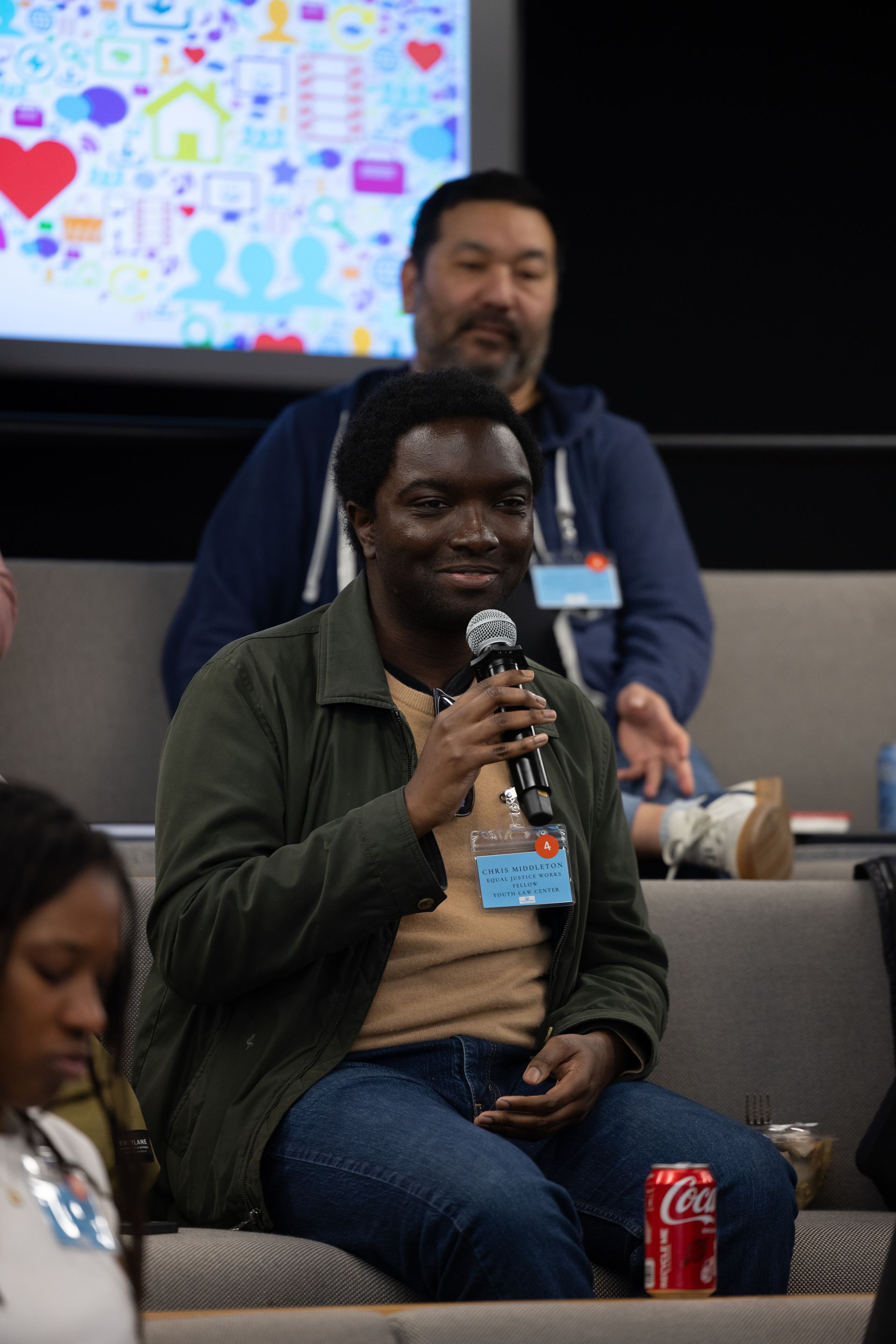 Audience member speaking into a microphone at a conference, with a slide screen behind them.