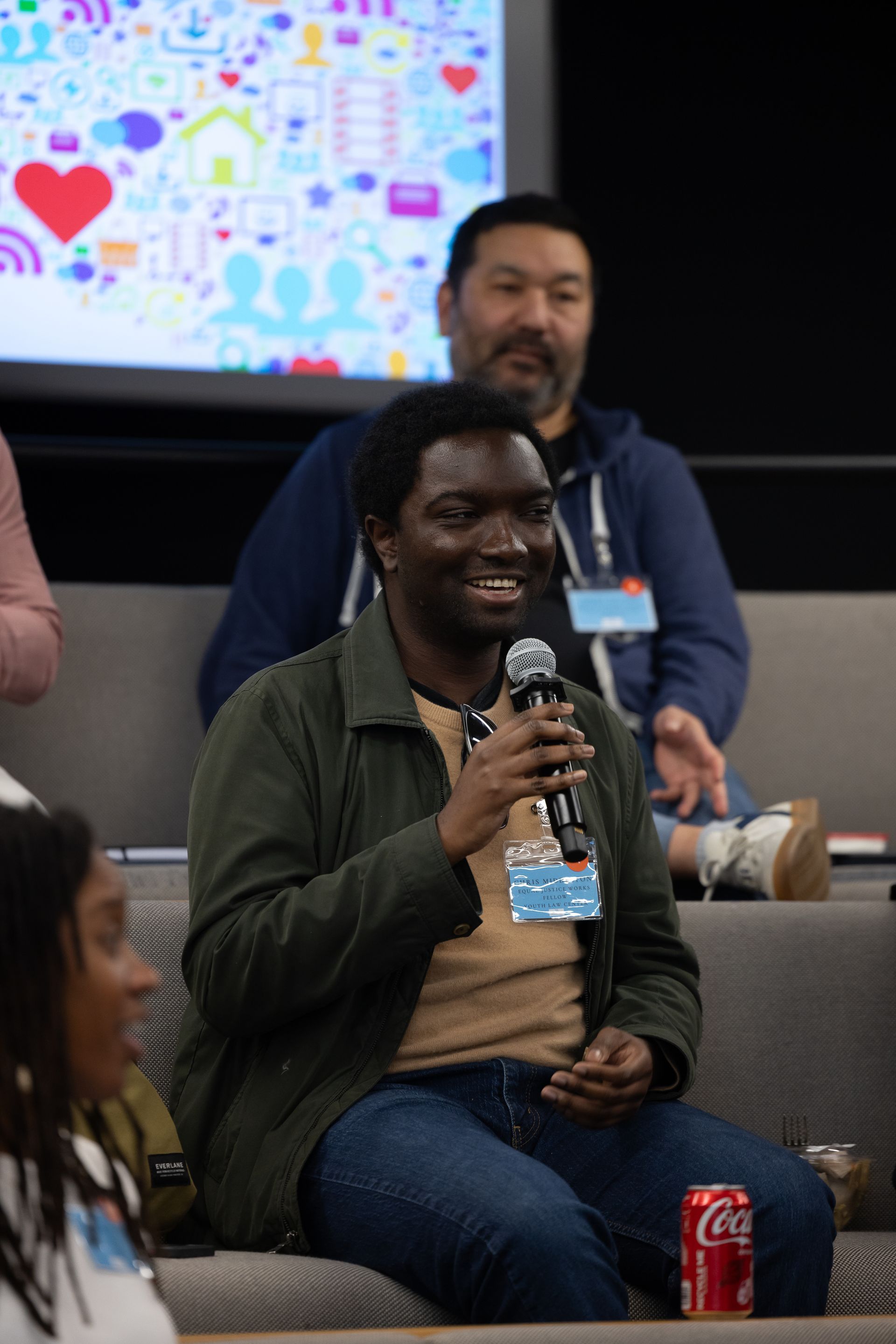 Audience member speaking into a microphone at a conference, seated in front of a presentation screen.