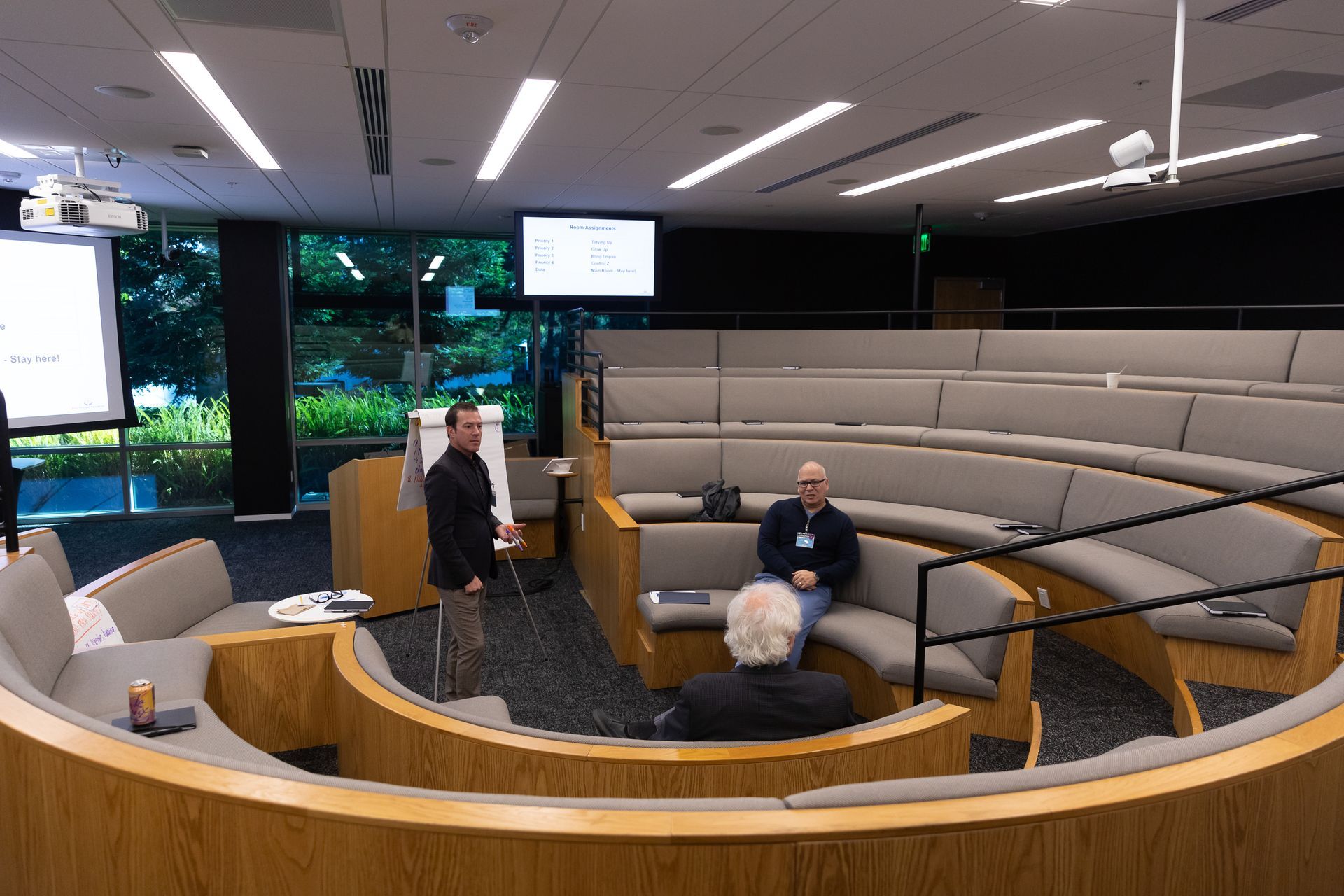 Curved lecture hall with three people seated and standing near podiums under large screens