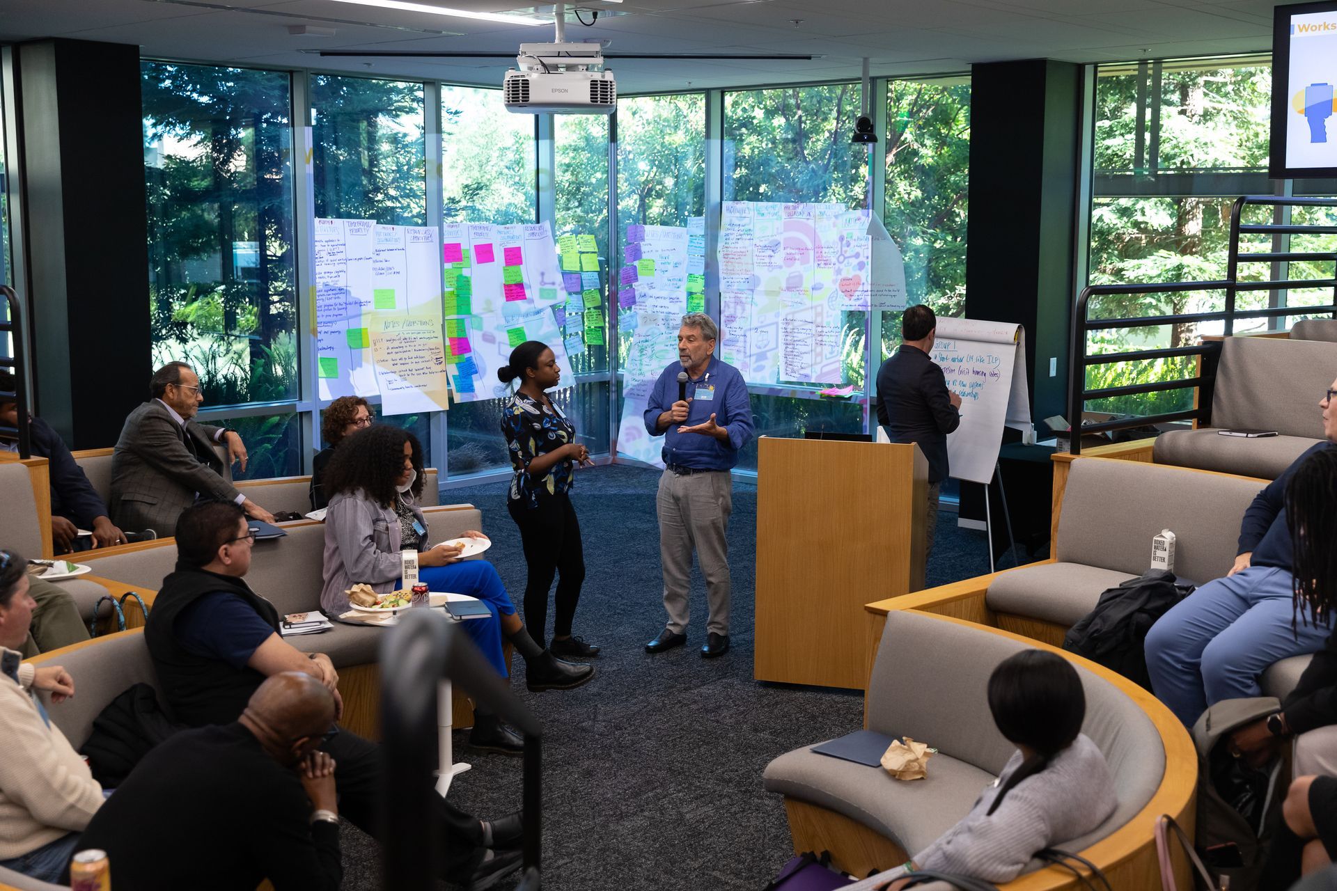 People gathered in a modern office lounge for a meeting, with speakers near a podium and windows behind them.