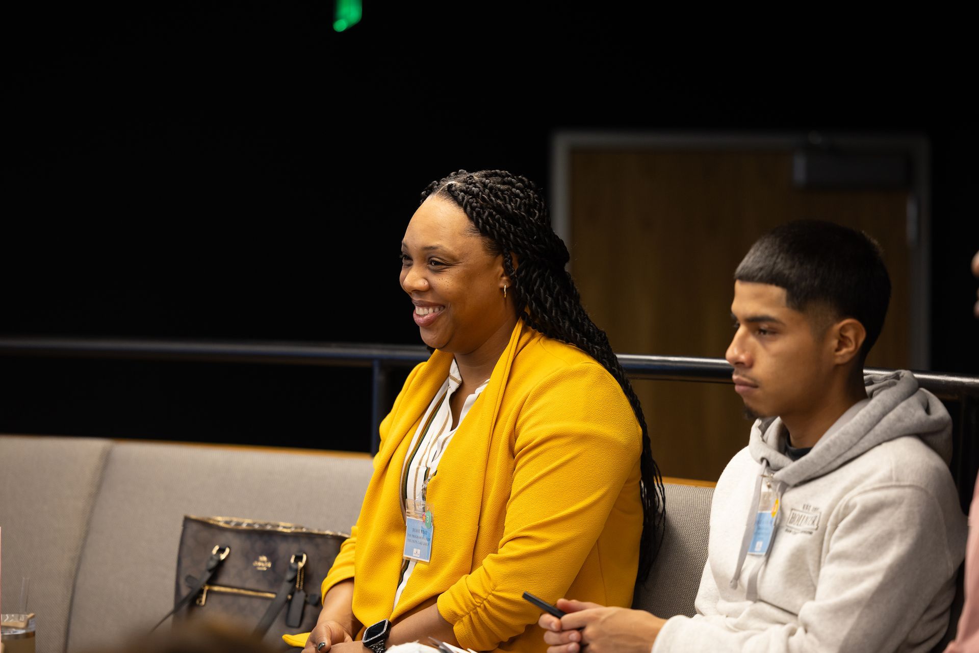 Two people seated indoors, one in a yellow blazer smiling and the other in a gray hoodie listening attentively.