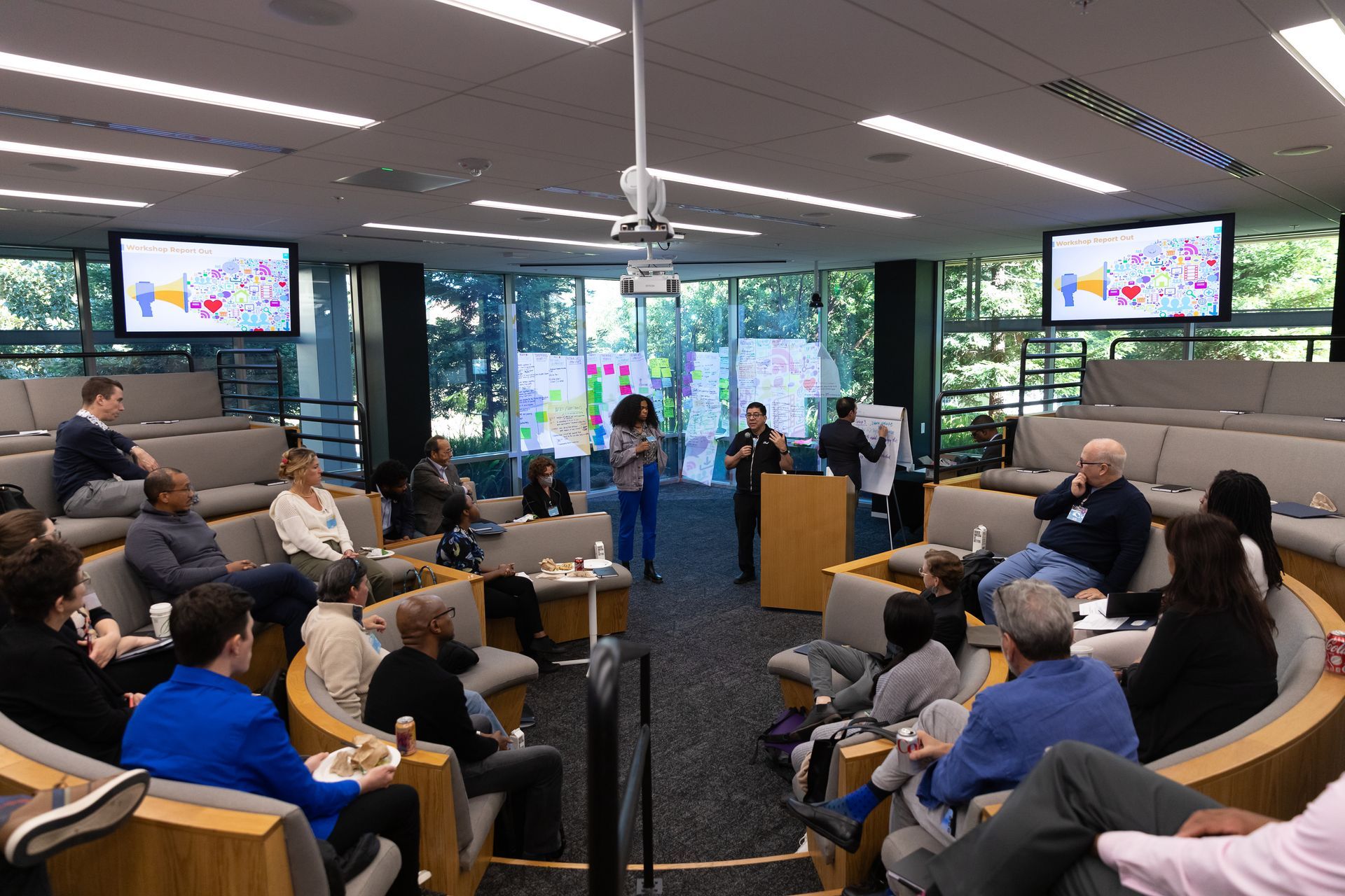 Conference panel in a bright room with seated audience and speakers at a podium, with screens displaying slides.