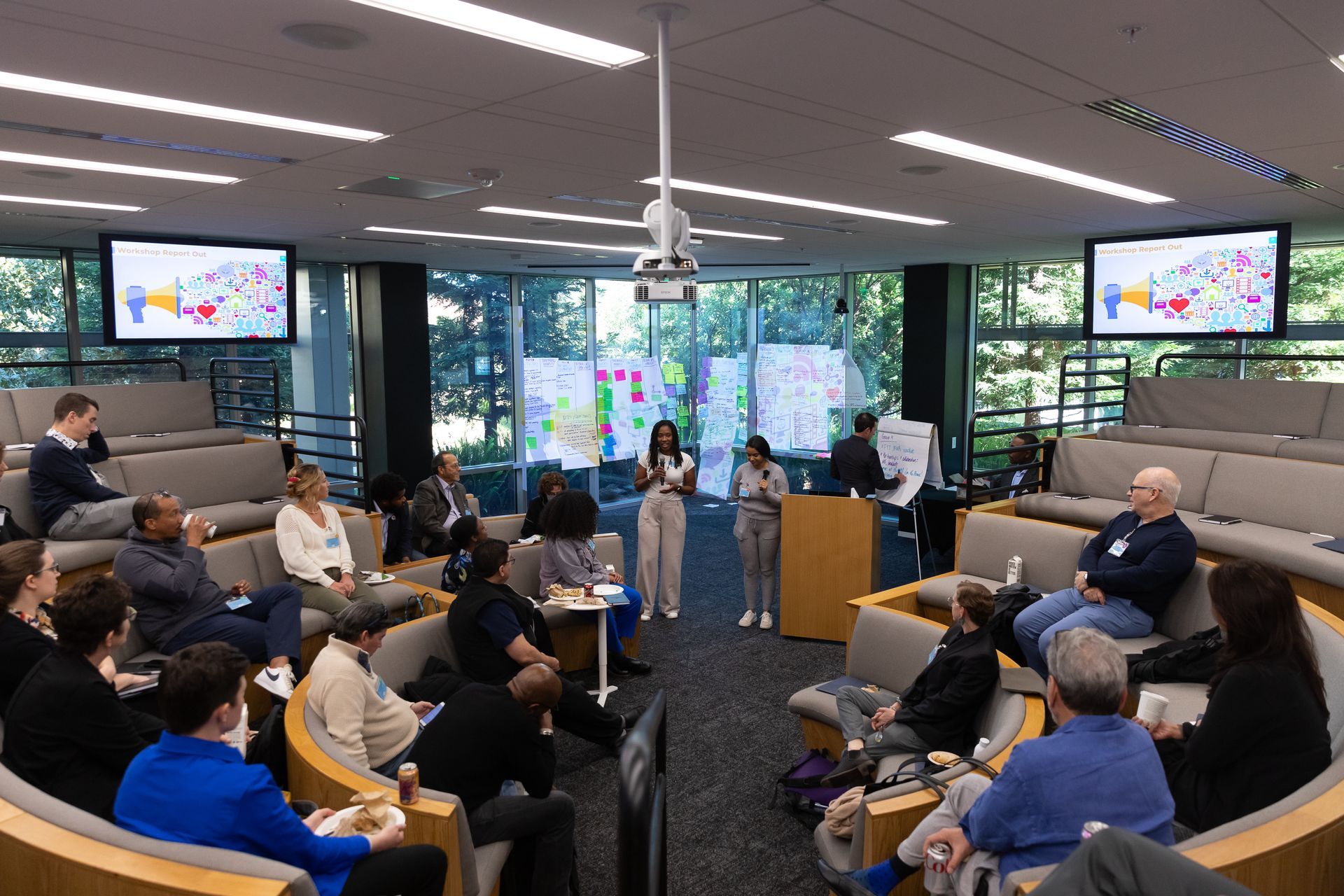 People seated in a conference room watching a speaker at a podium, with presentation screens on both sides.