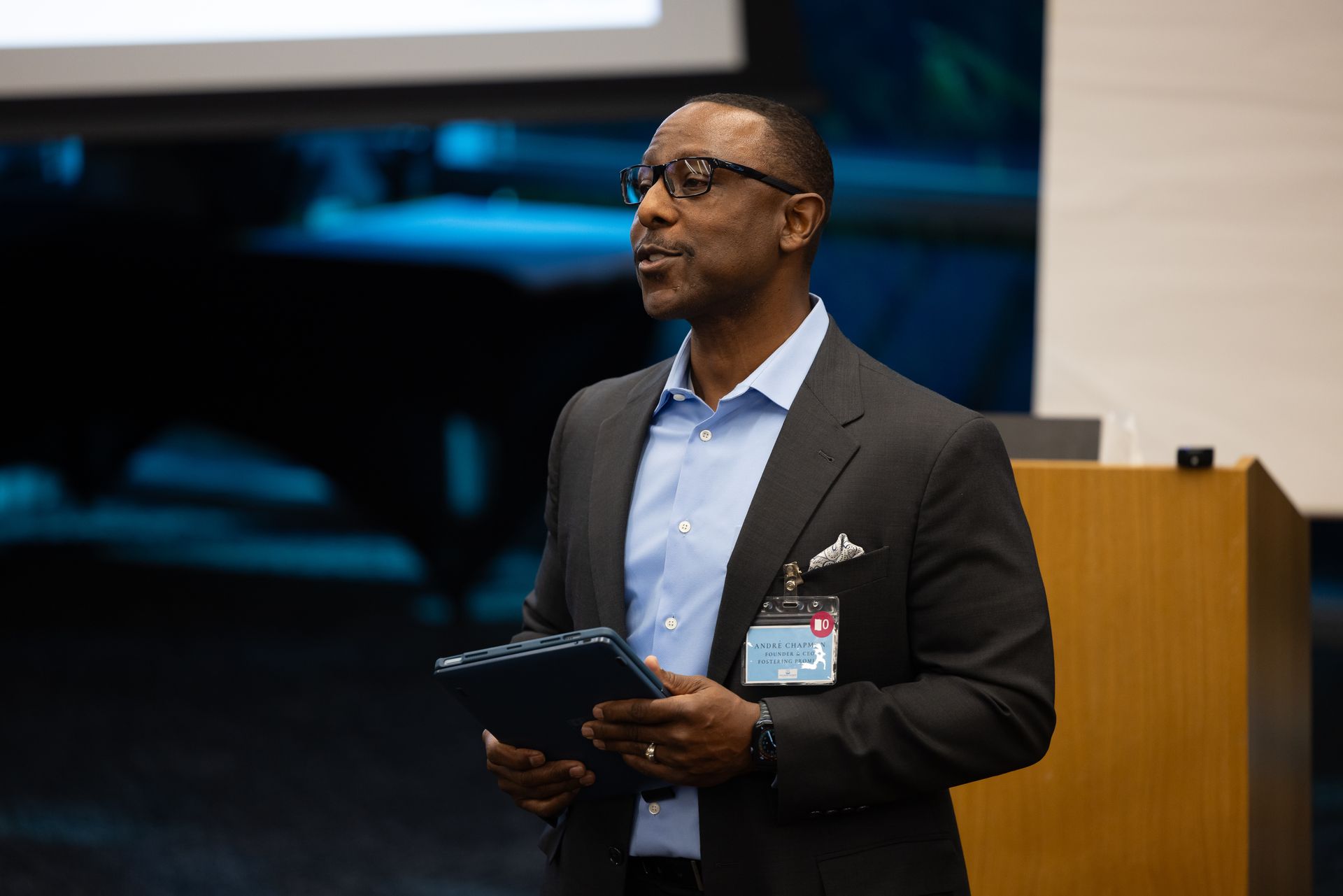 Man speaking at a podium, holding a tablet, in a conference room