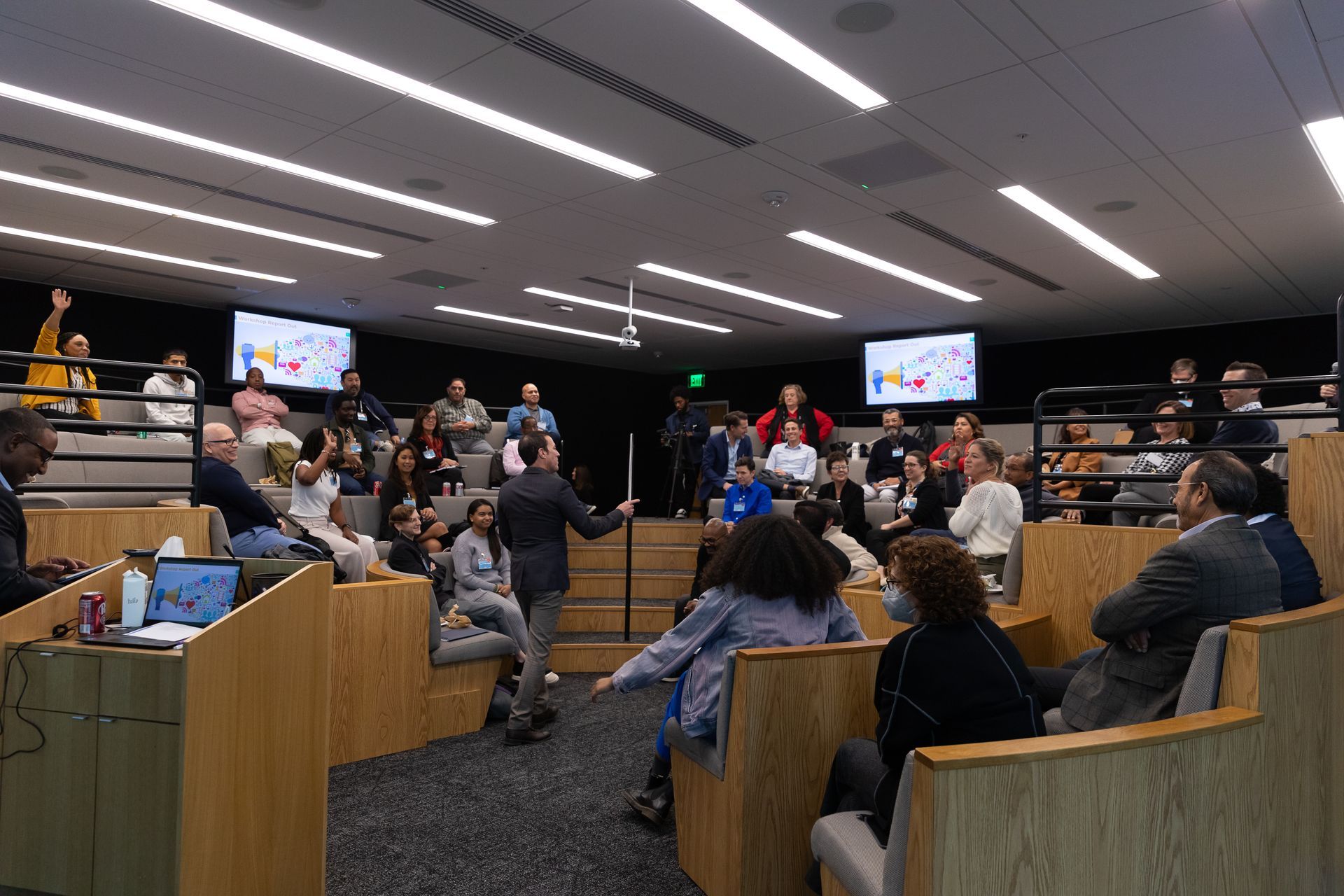 Meeting room with people seated in tiered rows, facing a presentation screen at the front.